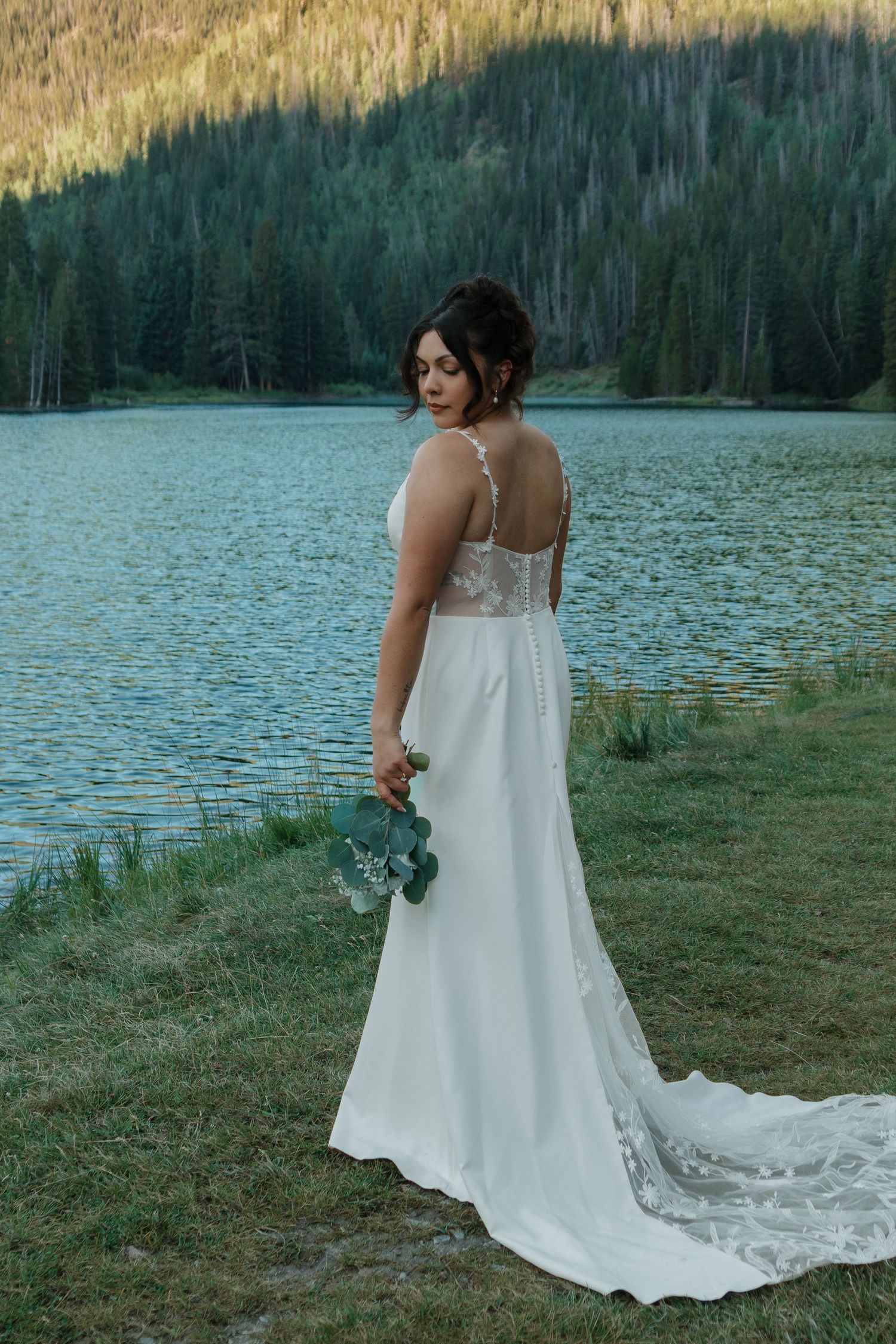 A white wedding dress flows elegantly against a lakeside mountain backdrop during golden hour.