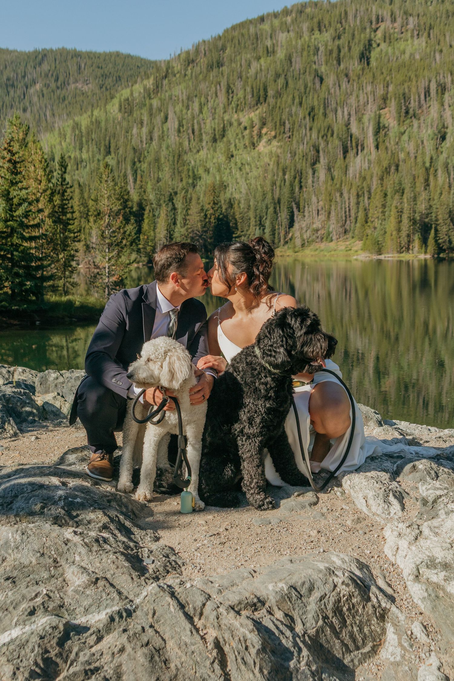 Wedding couple shares intimate moments on rocky shoreline with mountain lake vista.