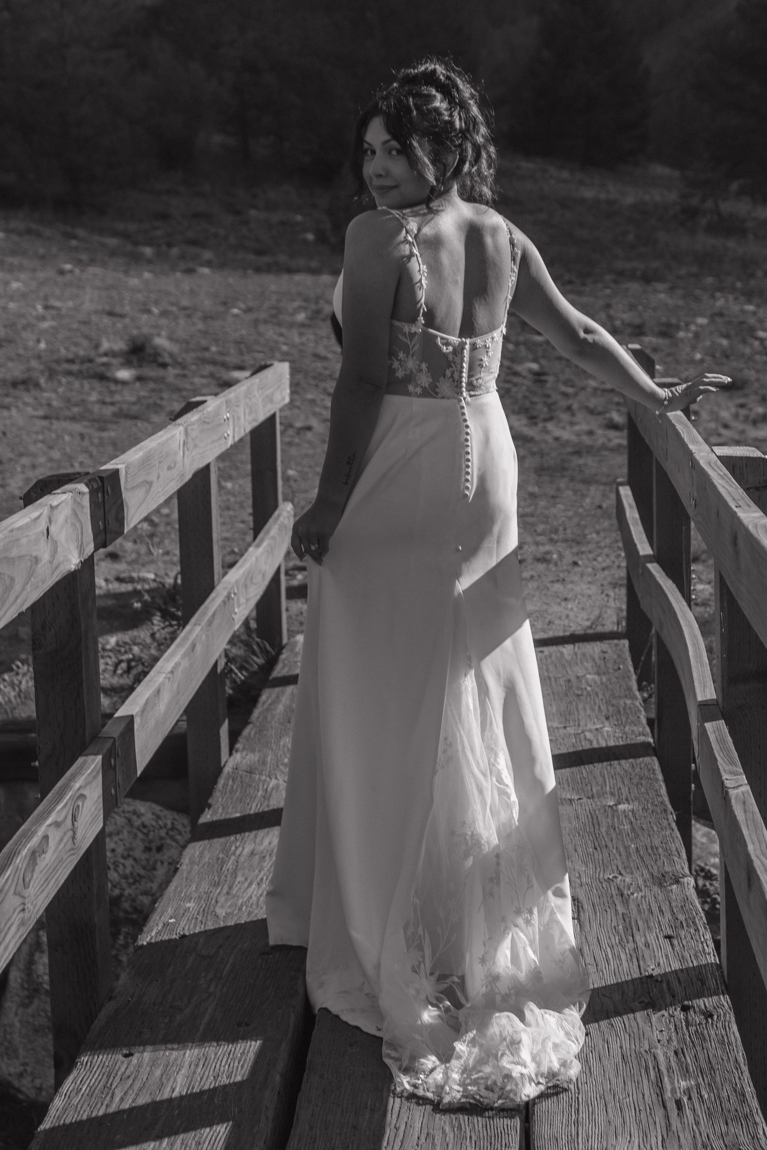 Black and white photo of person in wedding dress walking along wooden bridge railing.