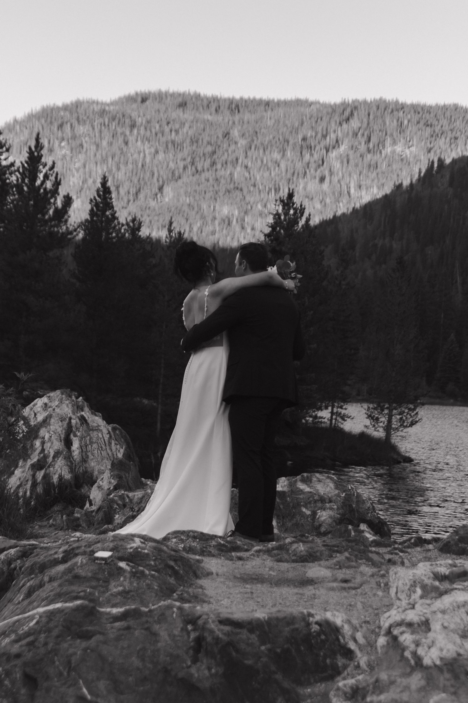 Silhouetted wedding couple stands on rocky outcrop near lake with mountain backdrop at dusk.
