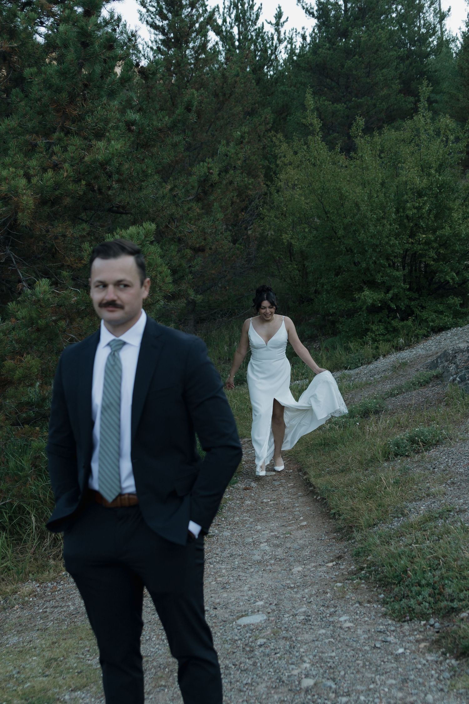 A couple walks down a scenic mountain path surrounded by evergreen trees at dusk.