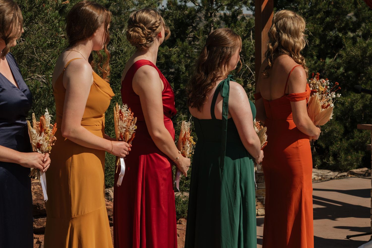 Wedding party members in colorful autumn-toned dresses standing in line outdoors.