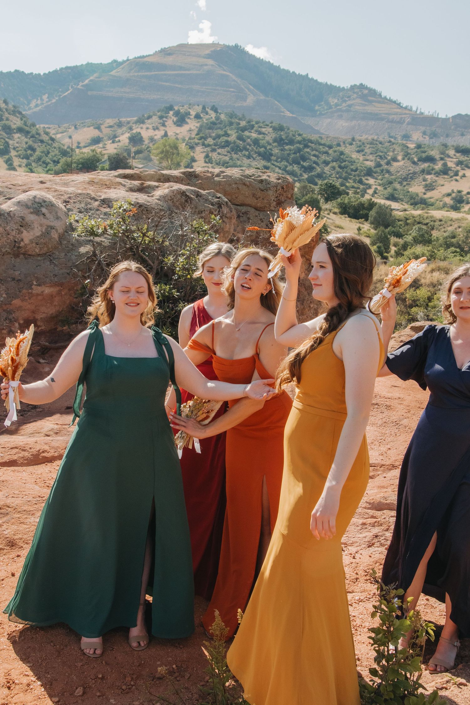 A bridal party in colorful dresses poses on red rocks with mountain views.