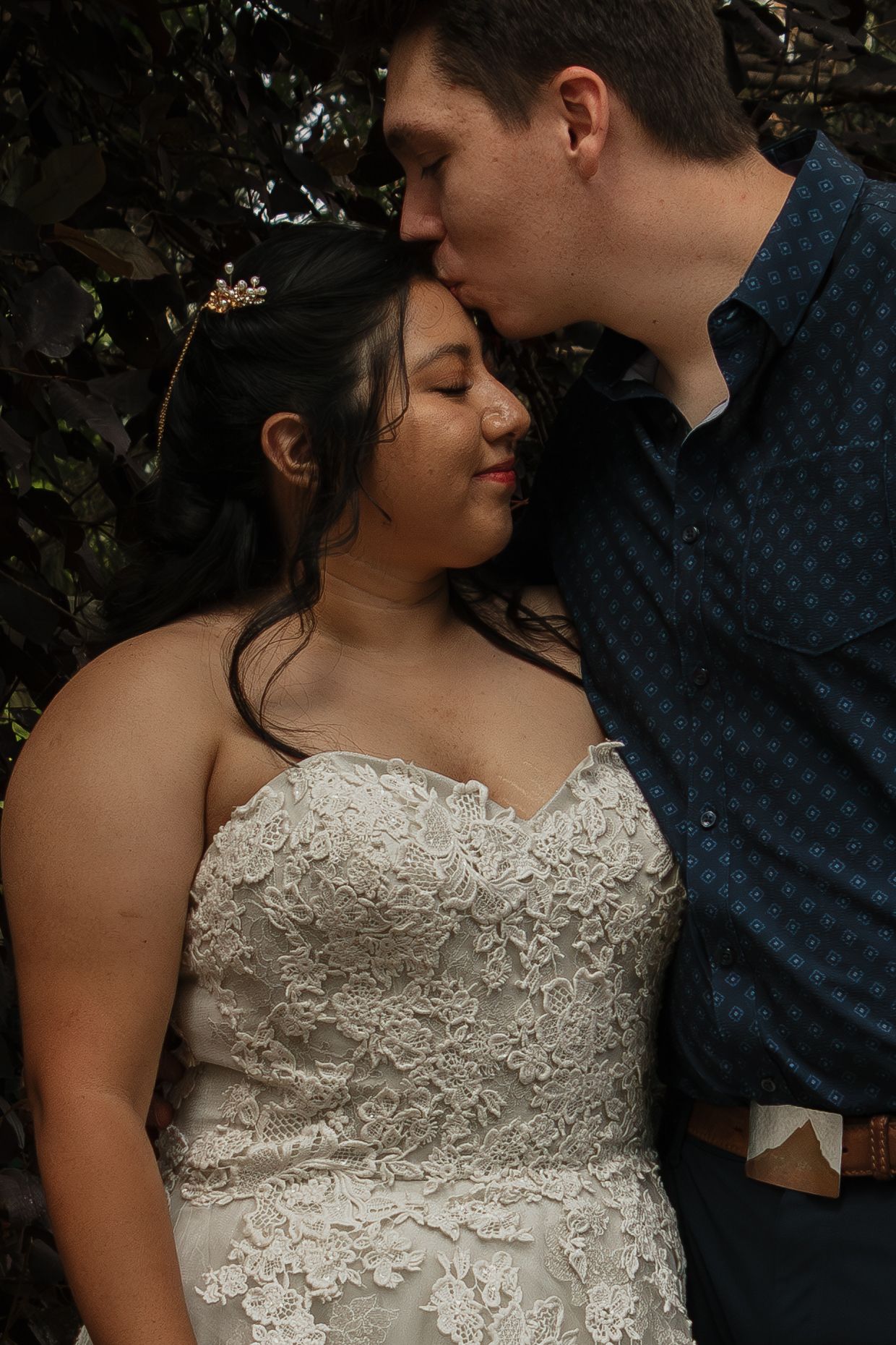 Intimate romantic moment shared between a couple, with bride wearing a detailed lace wedding dress against dark foliage.