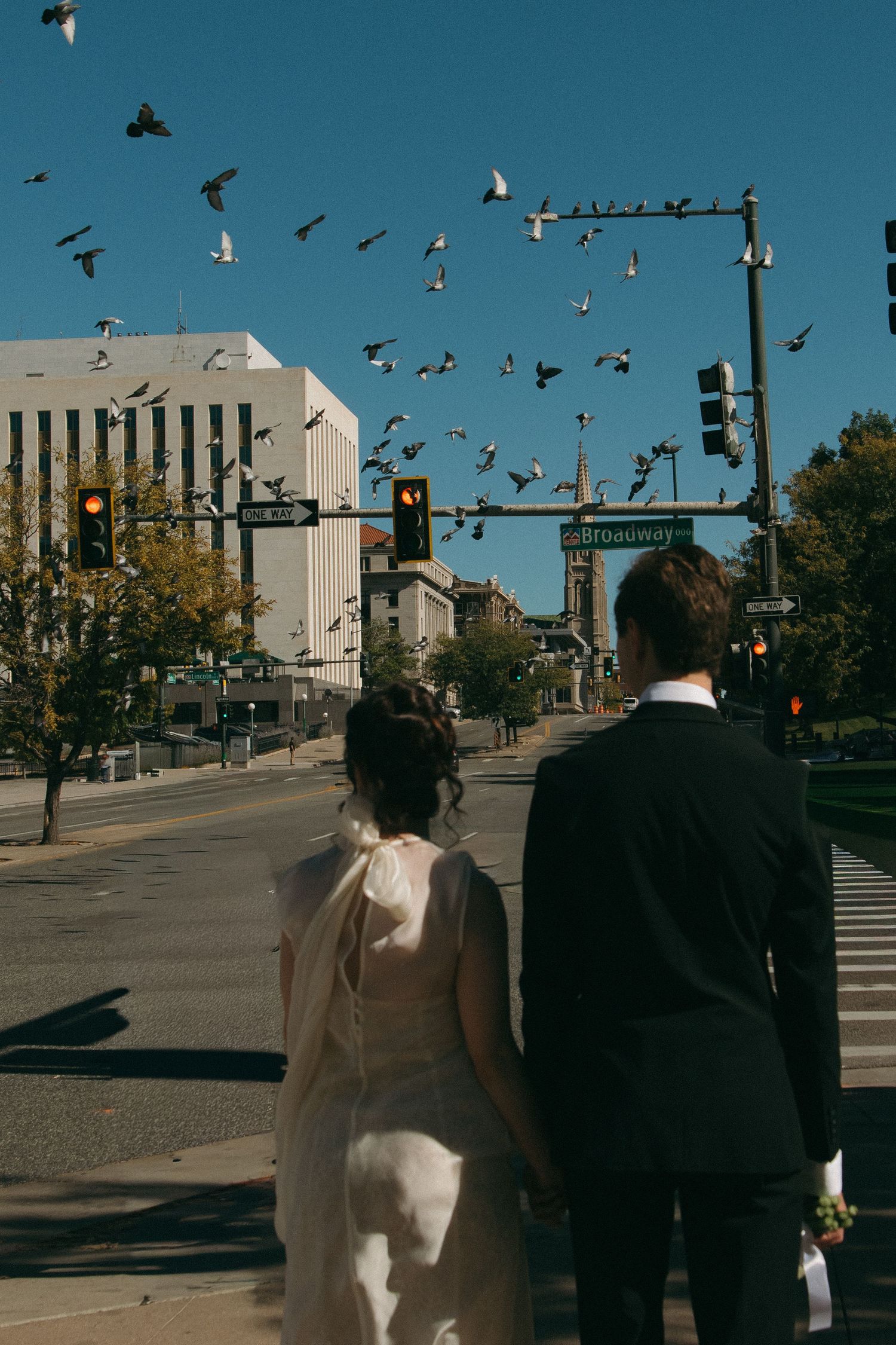 Couple walking on city street with pigeons flying overhead against blue sky.