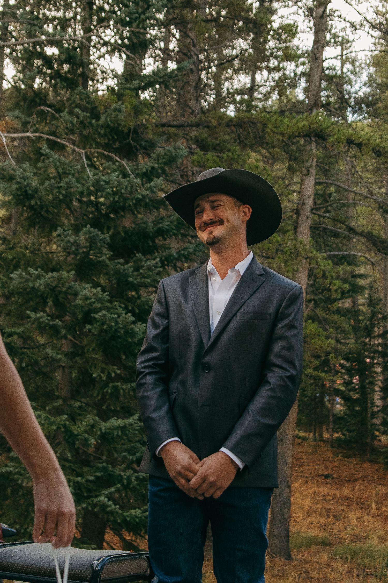 A groom in a black suit and cowboy hat stands in a wooded outdoor setting.