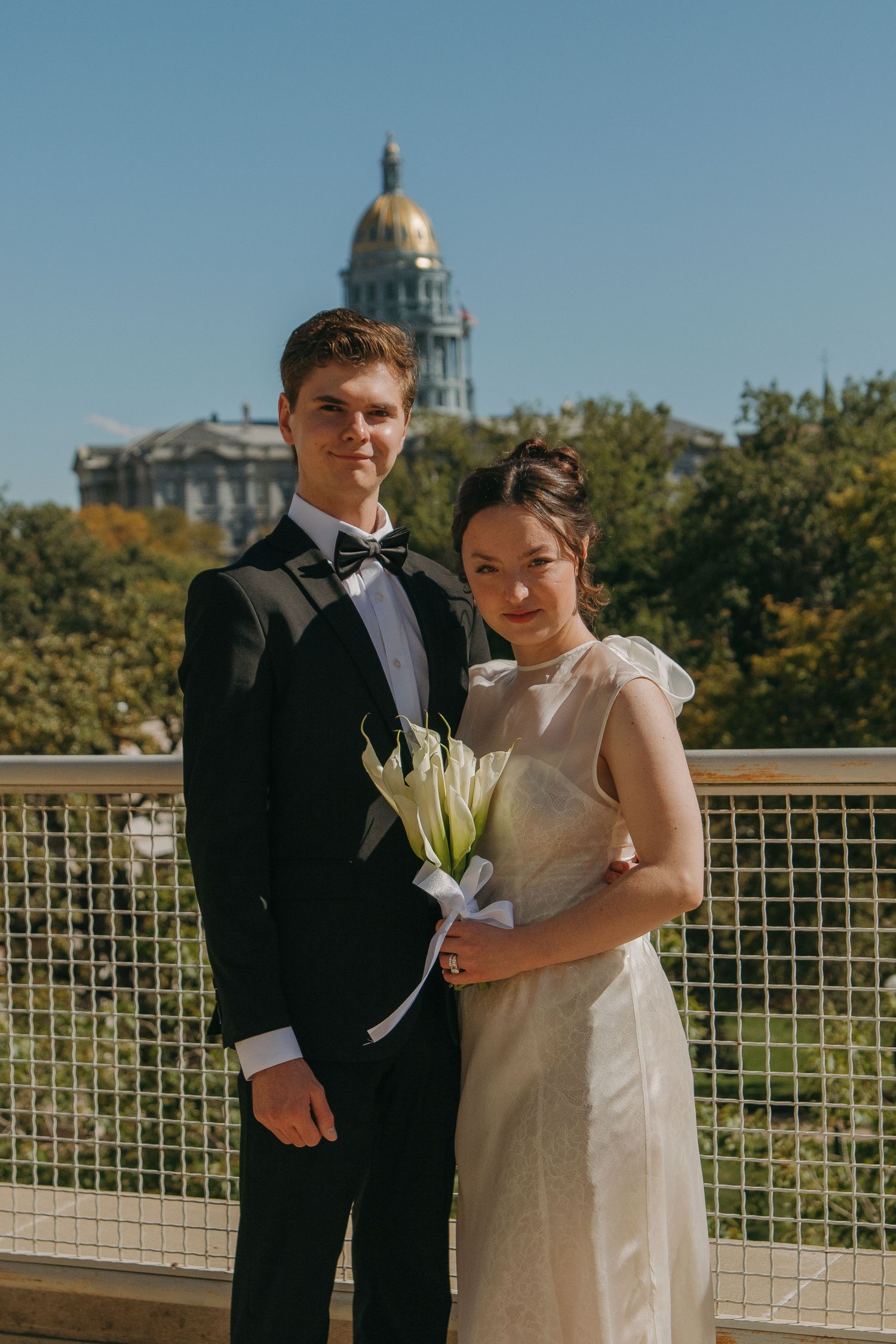 A couple in formal wedding attire poses together on an outdoor terrace with a domed building visible in the background.