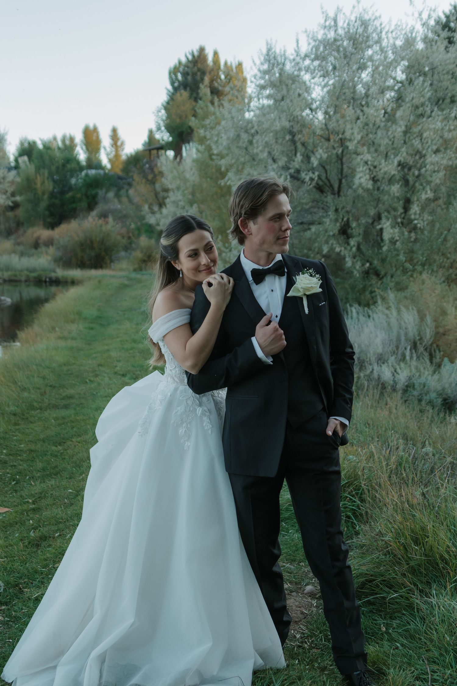 A couple in wedding attire poses together outdoors along a grassy path with trees in the background.