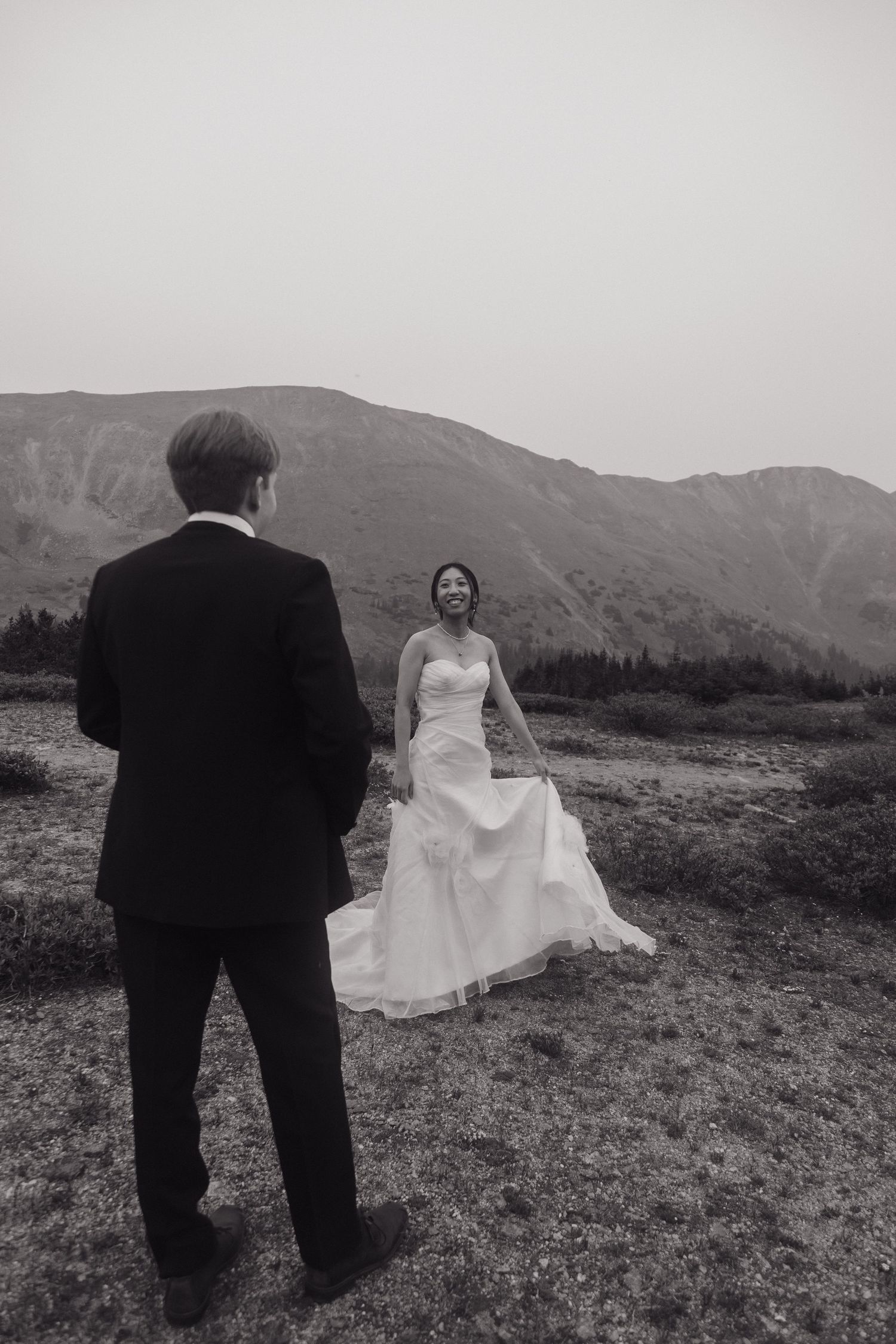 Black and white photo of bride lifting her gown walking toward groom on a mountain trail elopement.