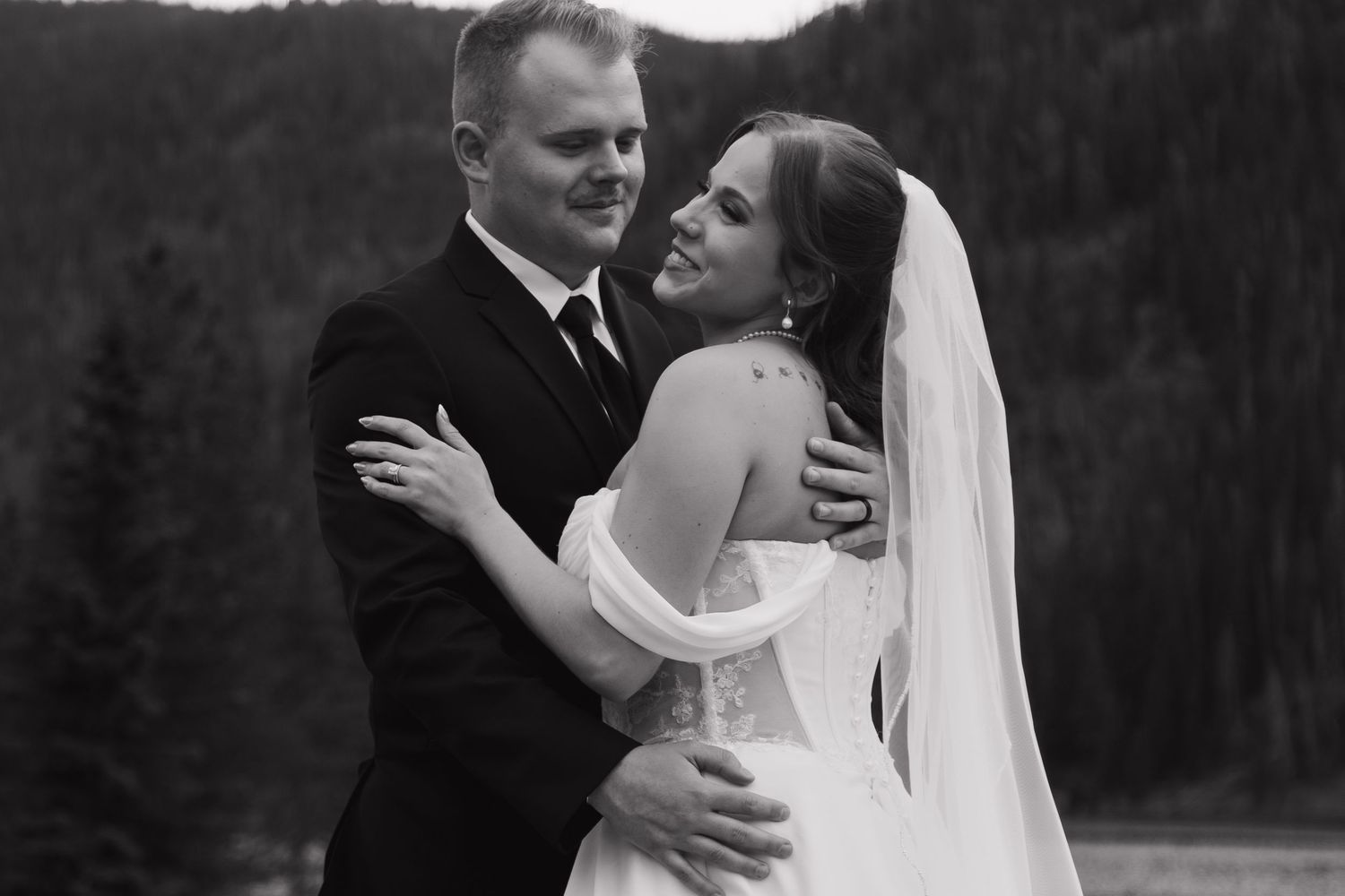 Black and white wedding photo of bride and groom embracing outdoors with forest backdrop.