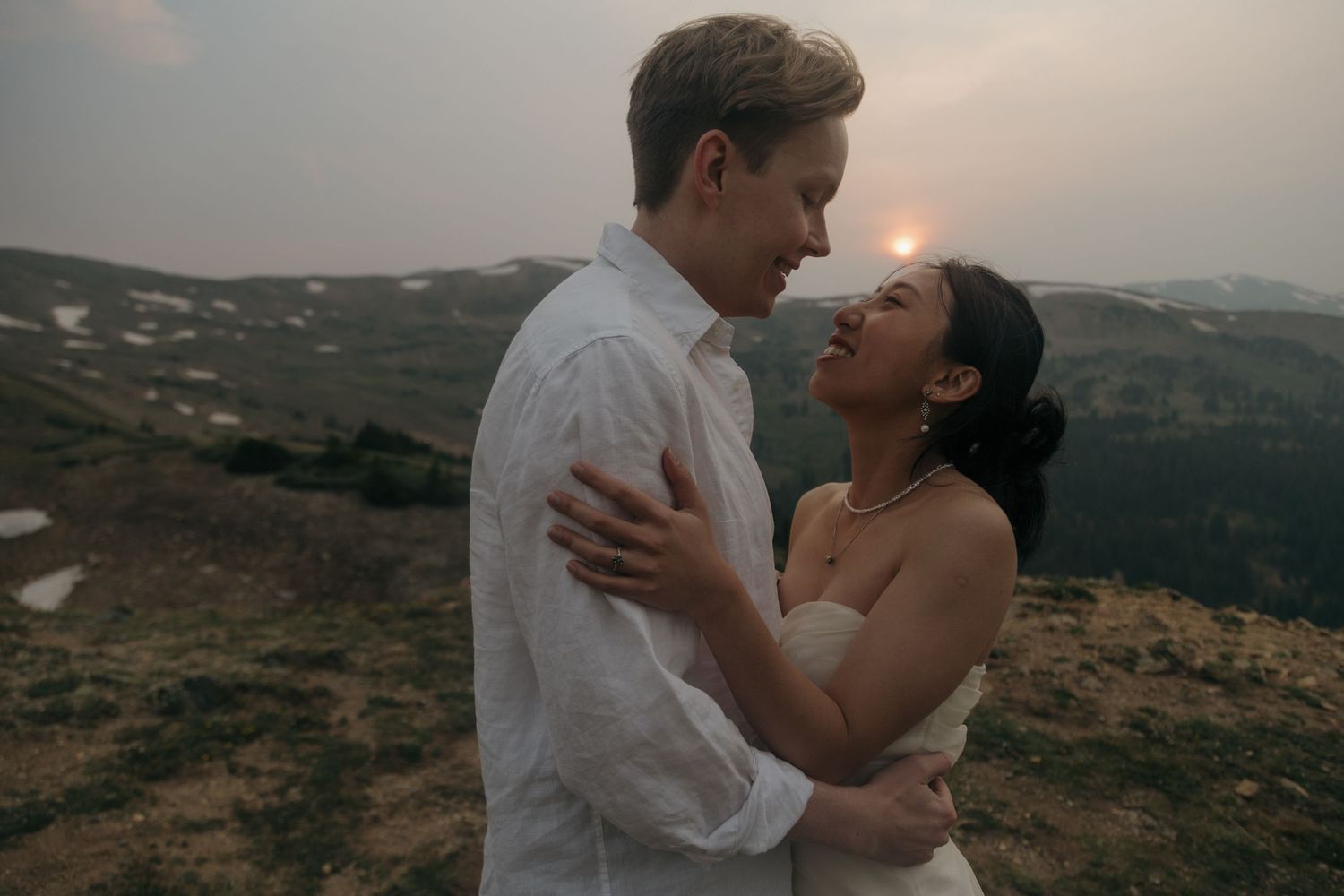 Couple in wedding attire embracing at golden sunset on an open mountain hillside with snow patches.