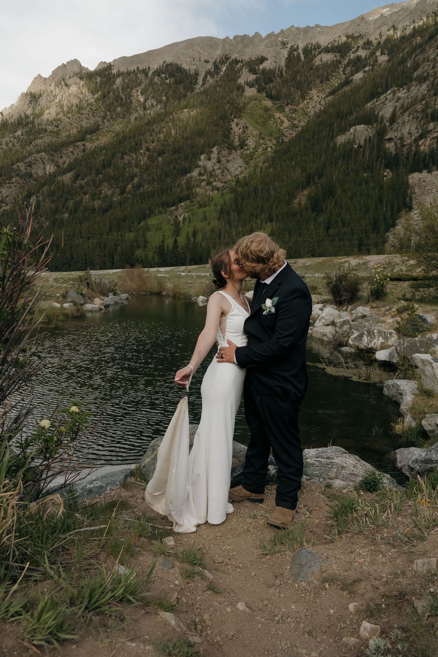 Bride and groom kiss beside a serene alpine lake with rocky mountains and evergreen trees behind them.
