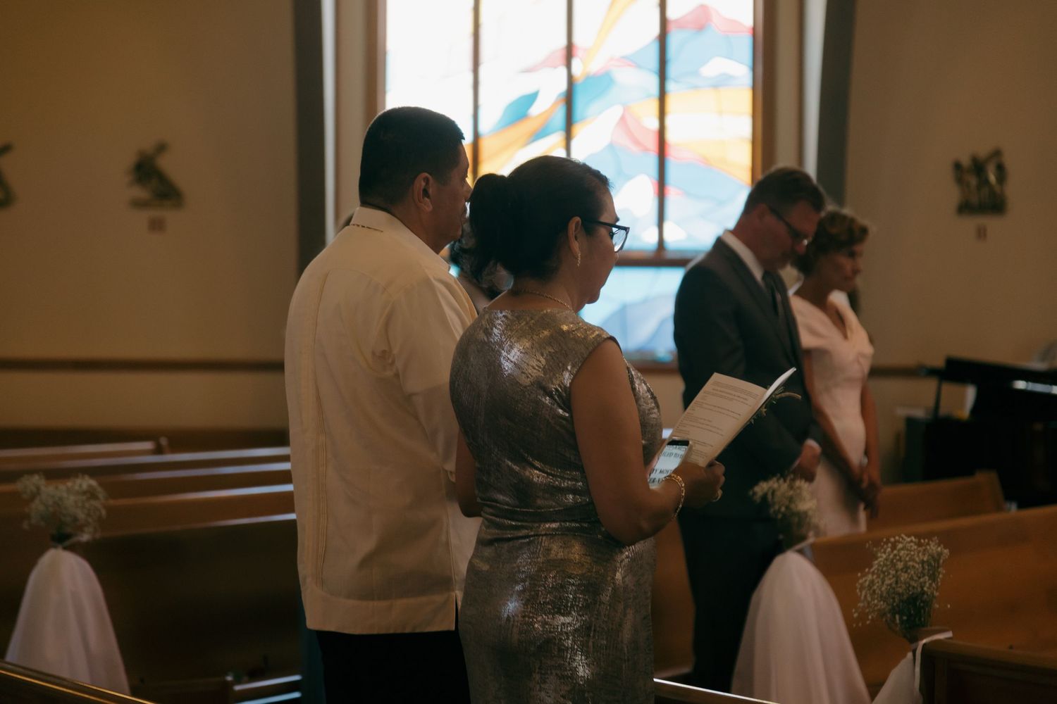 Wedding guests standing in church pews reading programs during a traditional wedding ceremony.