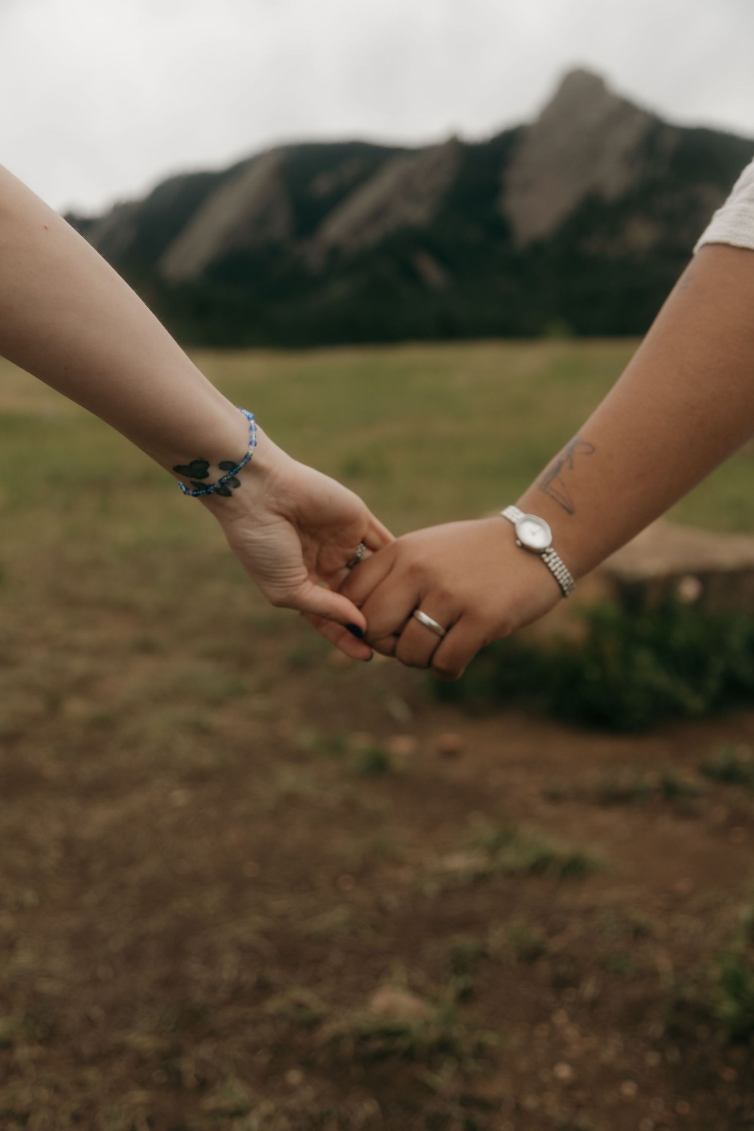 Close-up of two people holding hands with bracelets, rings, and a watch, with mountain landscape behind.