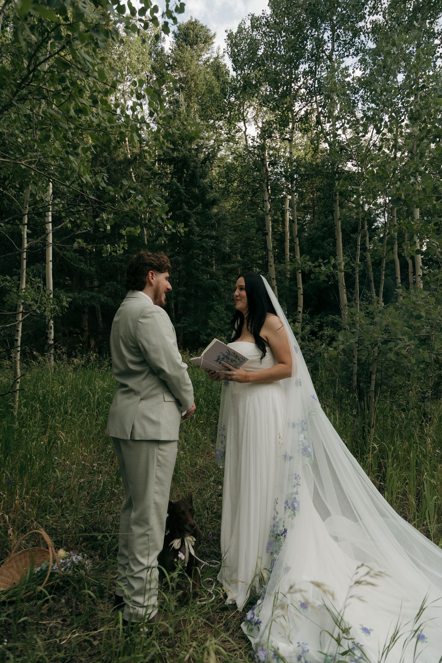 Bride in floral veil reads vows to groom in light gray suit during intimate forest elopement ceremony.
