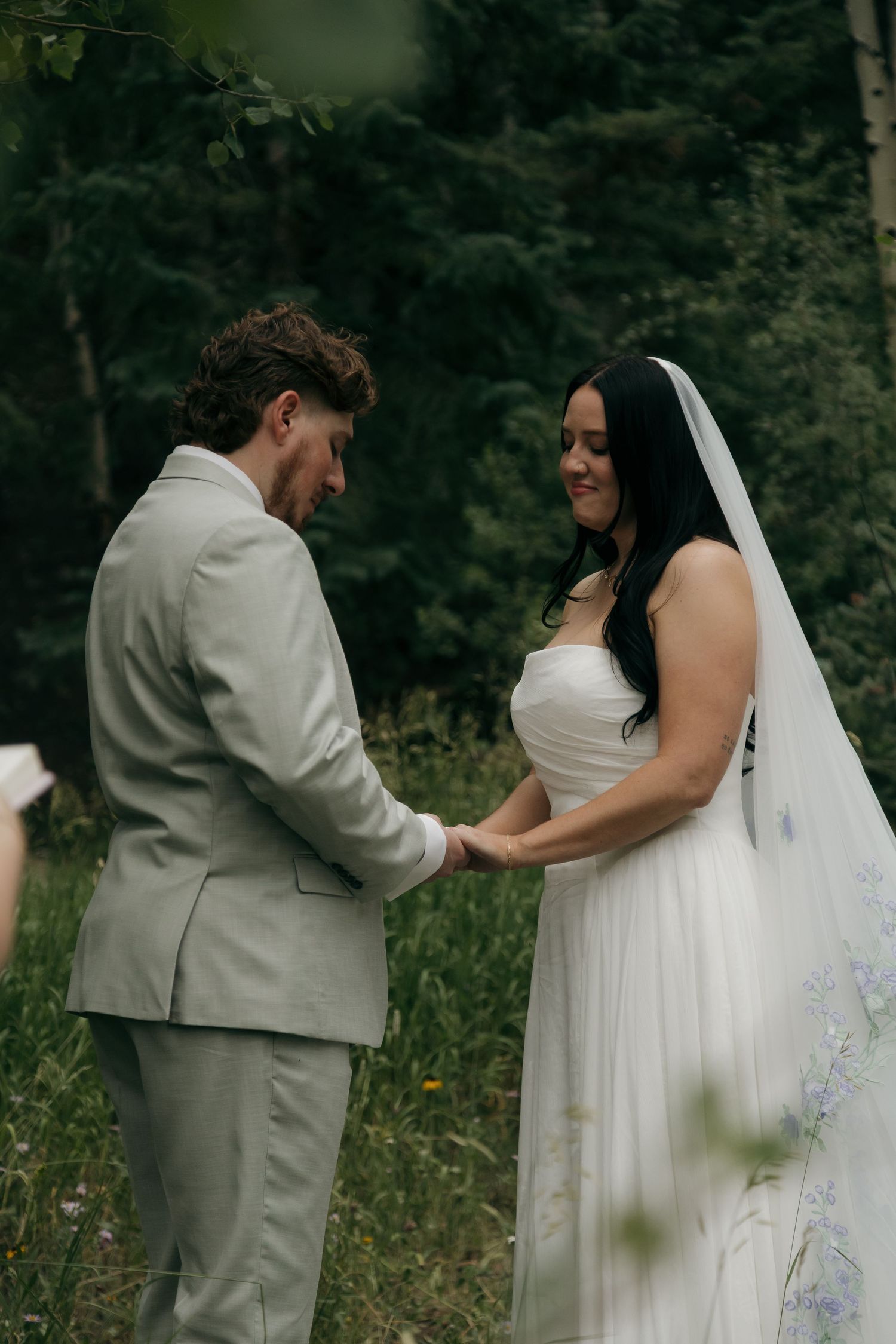 Bride and groom holding hands during outdoor forest wedding ceremony, bride in white gown with veil, groom in light gray suit.