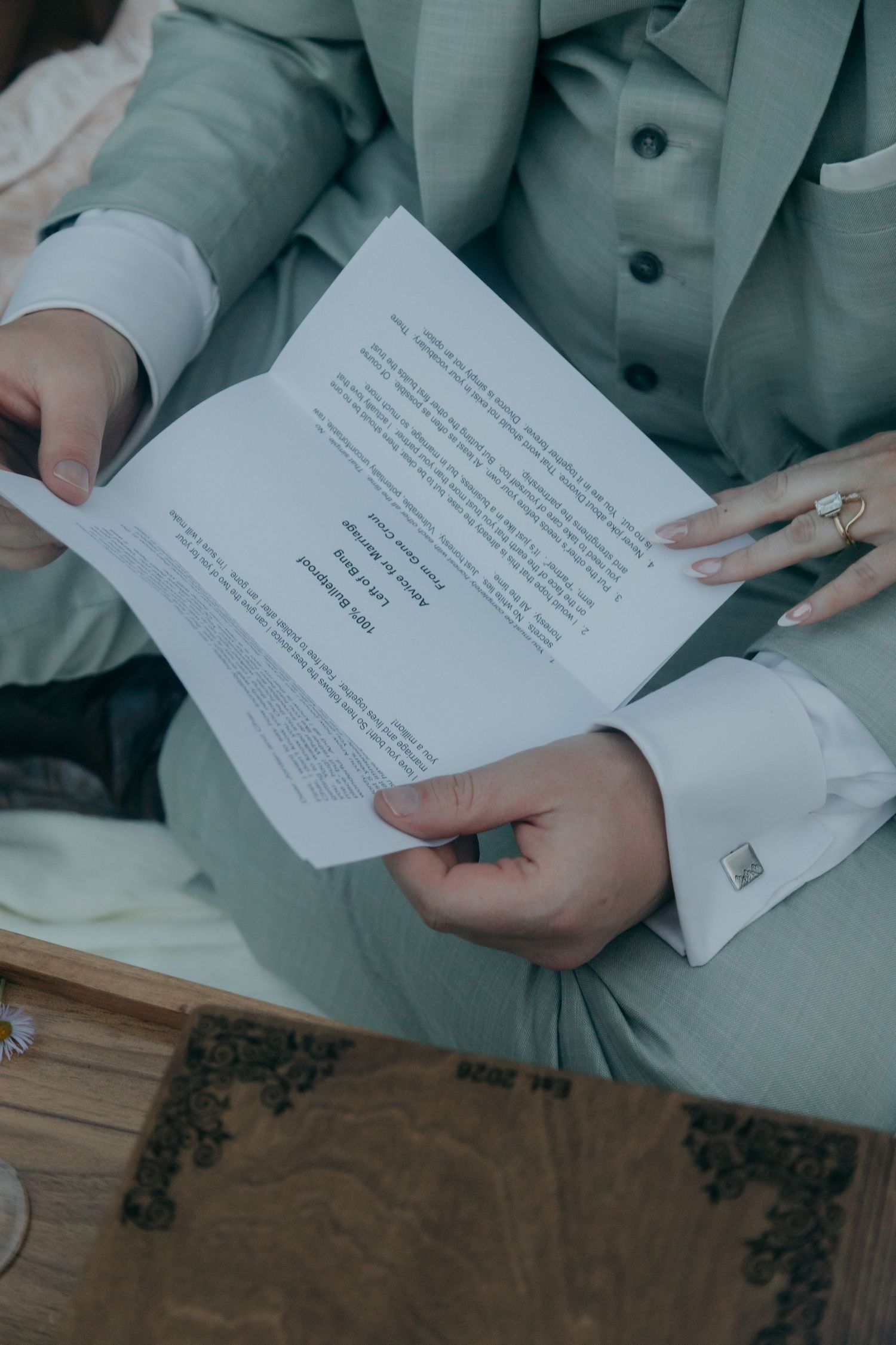 Close-up of groom in grey suit reading wedding vows document with manicured nails visible.