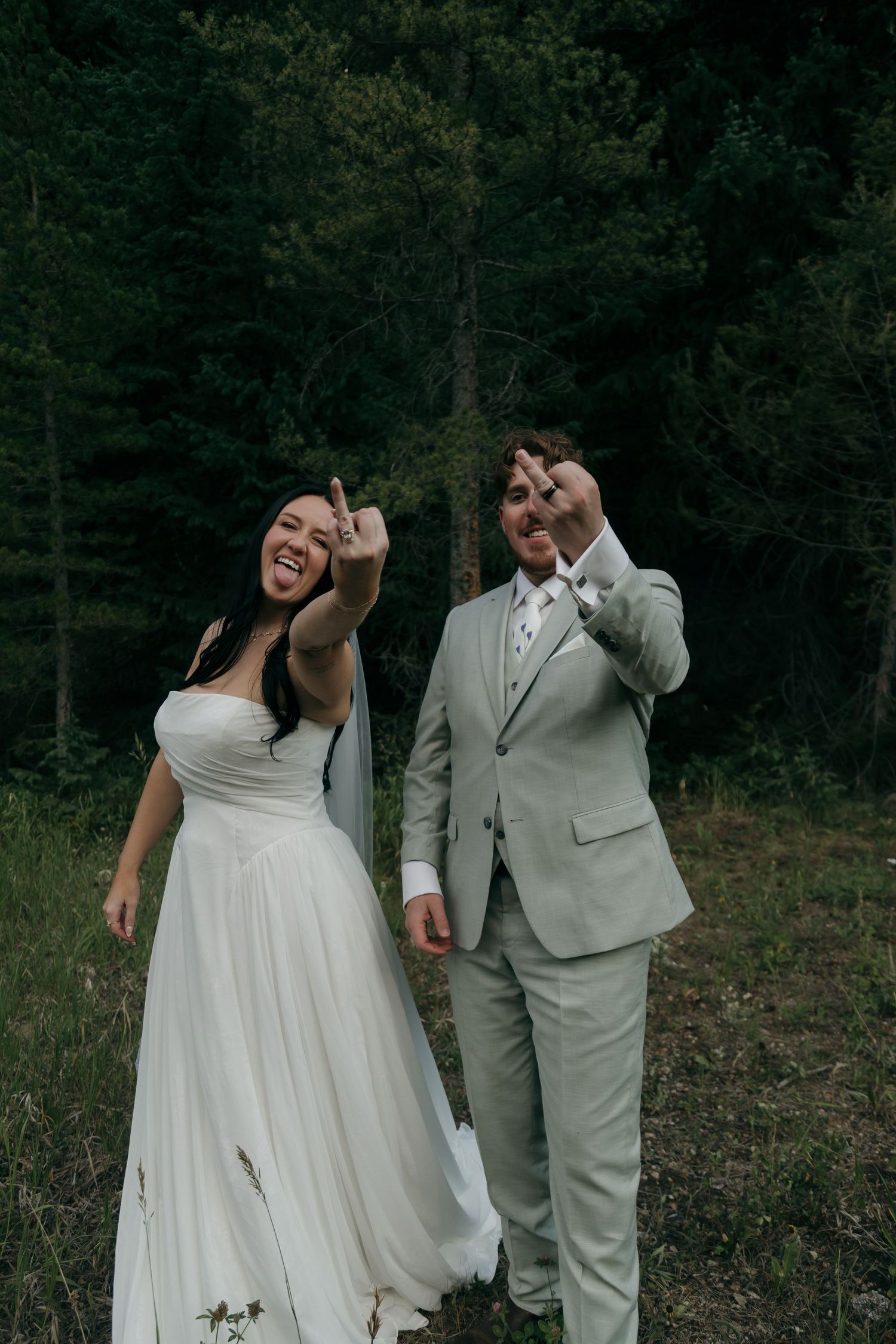 Laughing bride and groom in forest setting flipping off camera playfully in wedding attire.