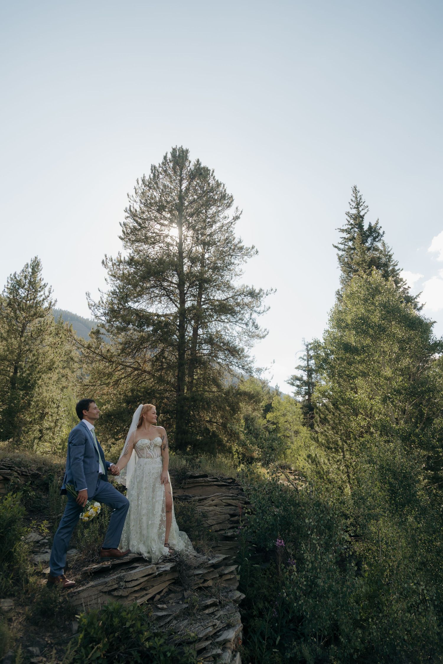 Bride and groom holding hands on a rocky ledge surrounded by tall pine trees in bright sunlight.