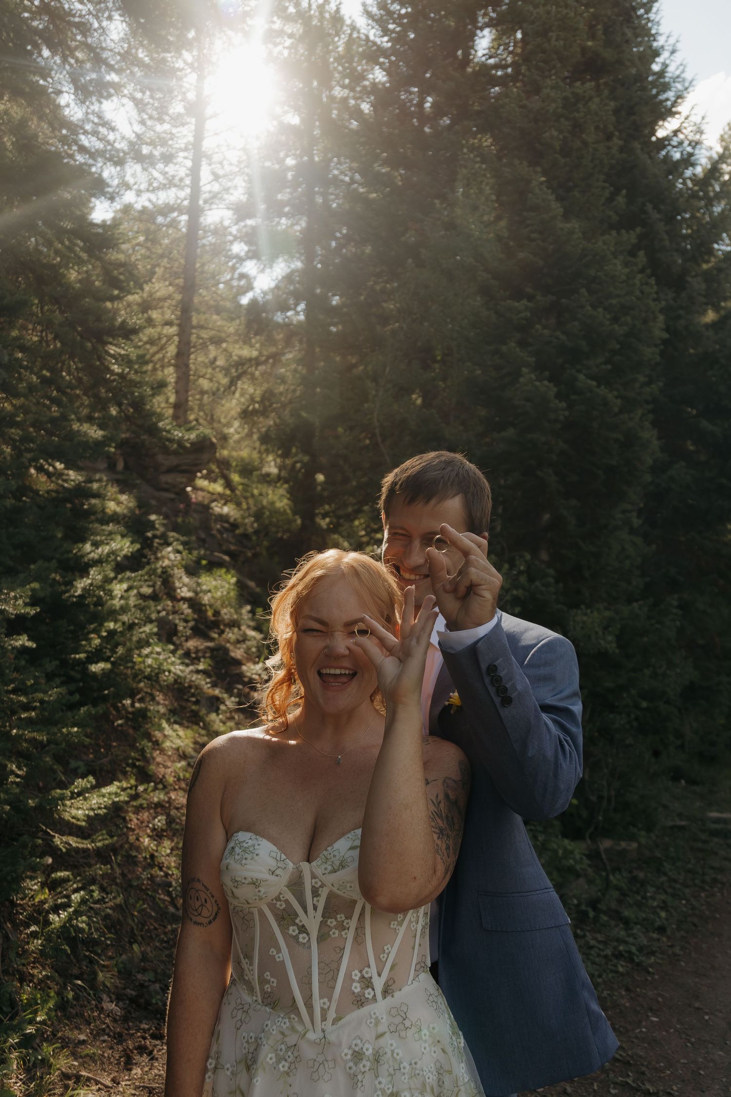 Playful bride and groom making silly faces in sunlit forest during wedding photo session.