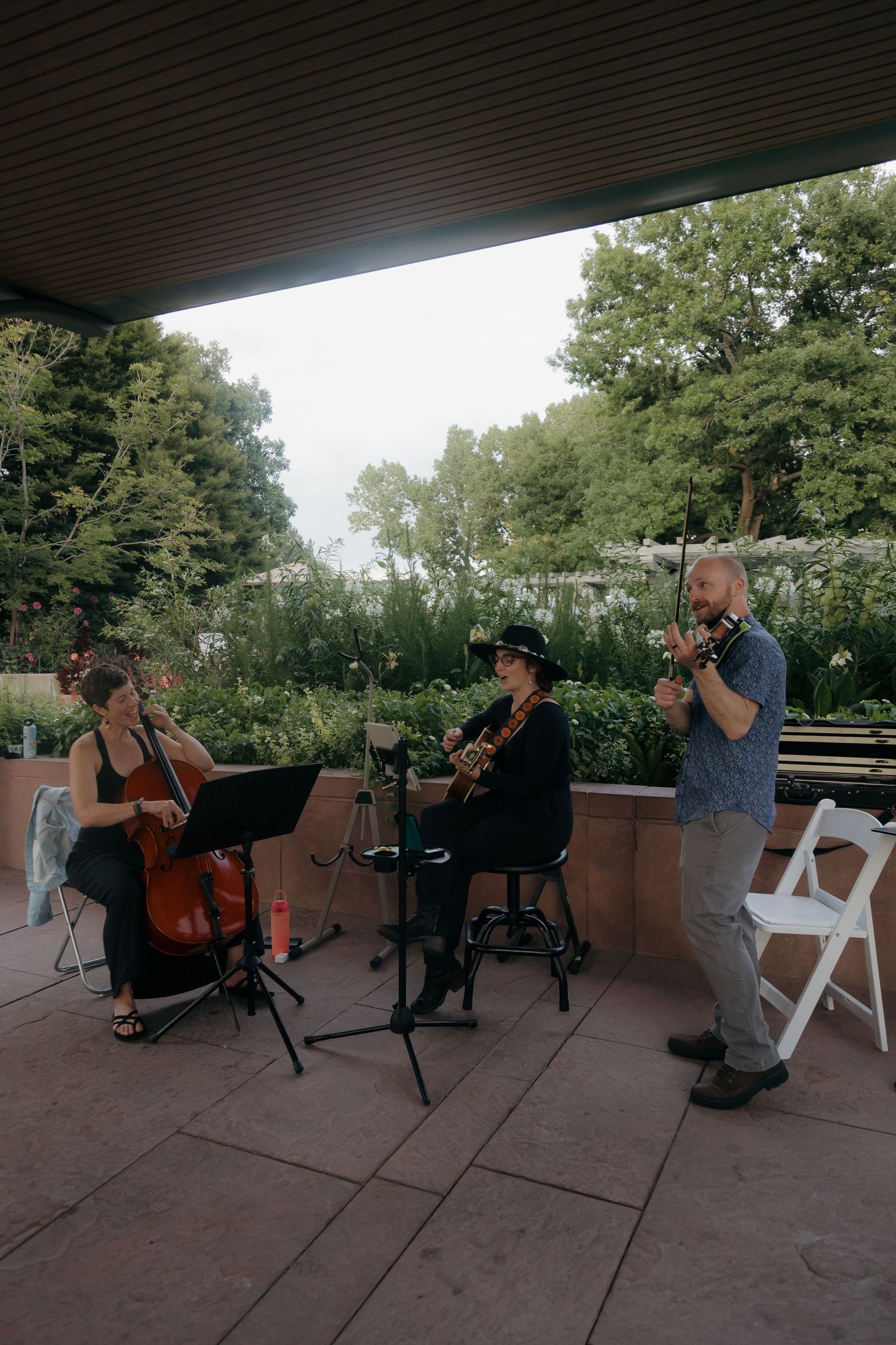 String trio performs outdoors at a wedding ceremony on a patio surrounded by lush green trees.