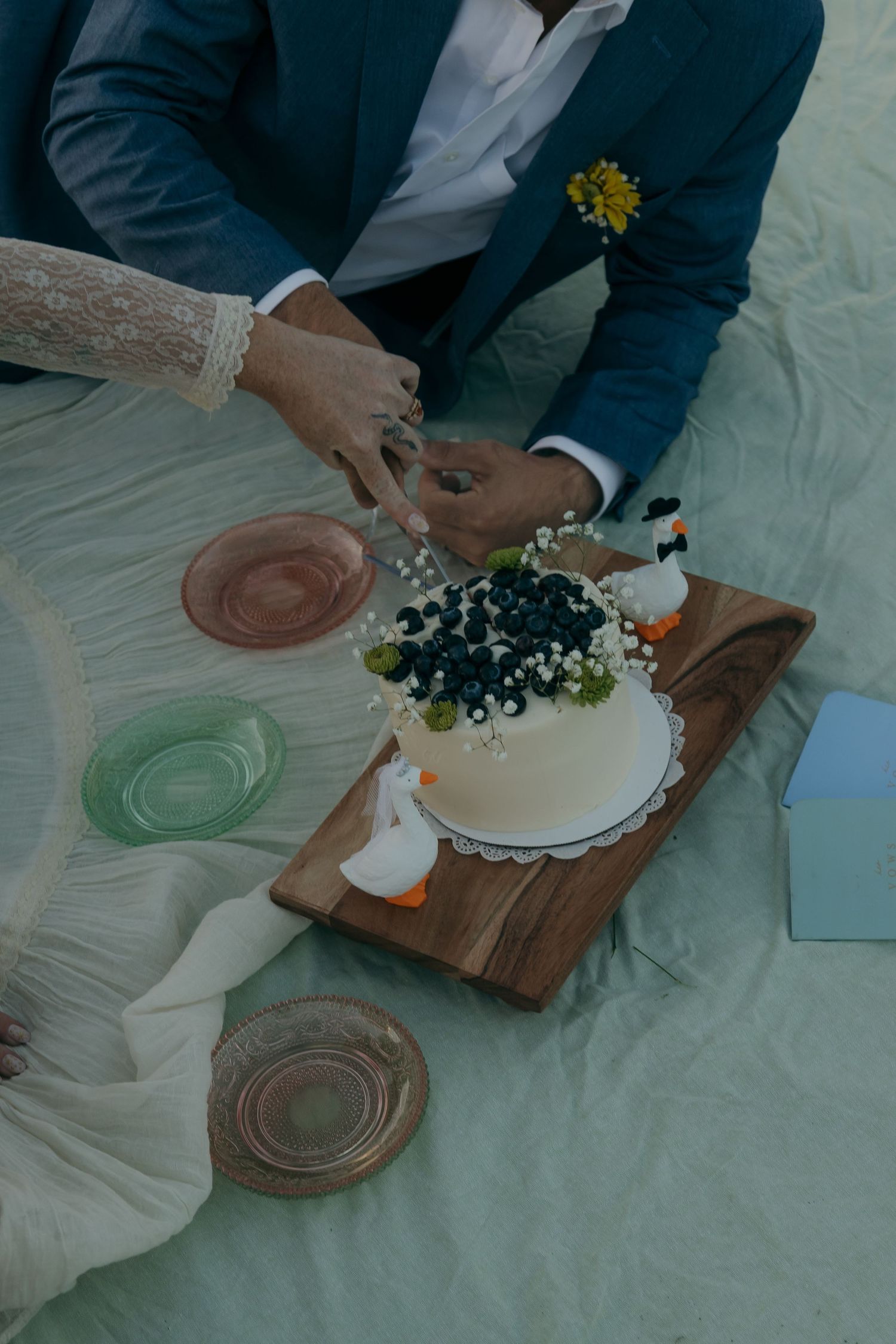 Newlyweds cutting a white floral wedding cake on a wooden board surrounded by colorful vintage glass plates.