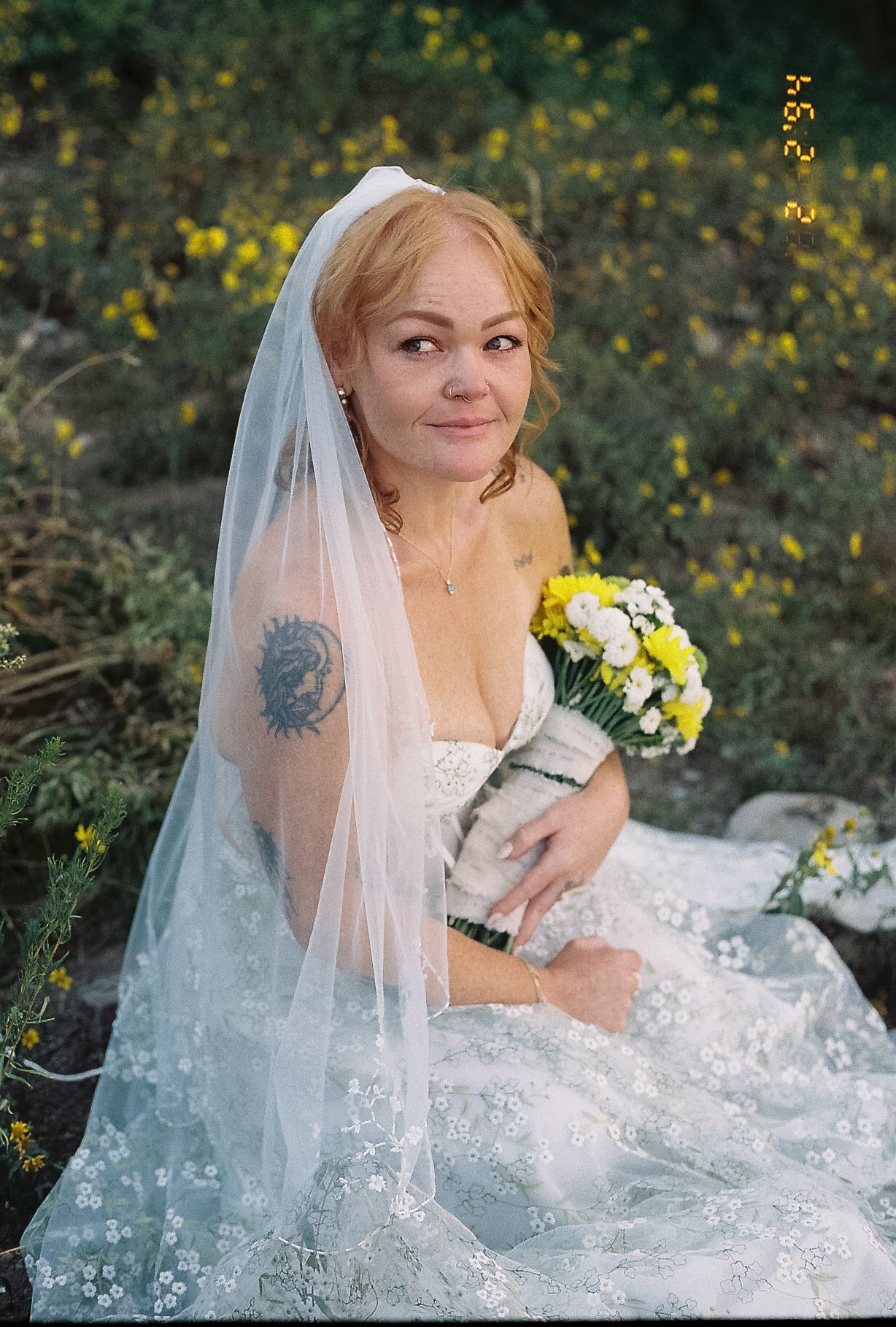 Smiling bride in lace gown and veil holding yellow and white bouquet seated among yellow wildflowers.
