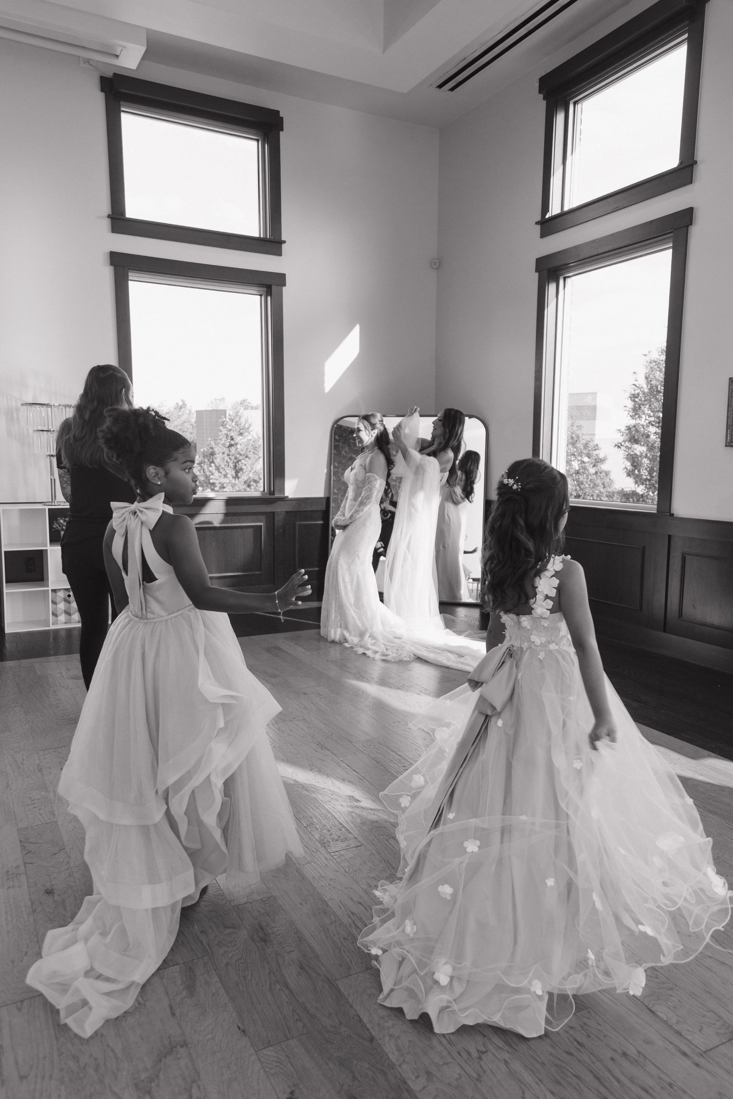 Two flower girls in white dresses hold a bride's train before a mirror in a sunlit room.