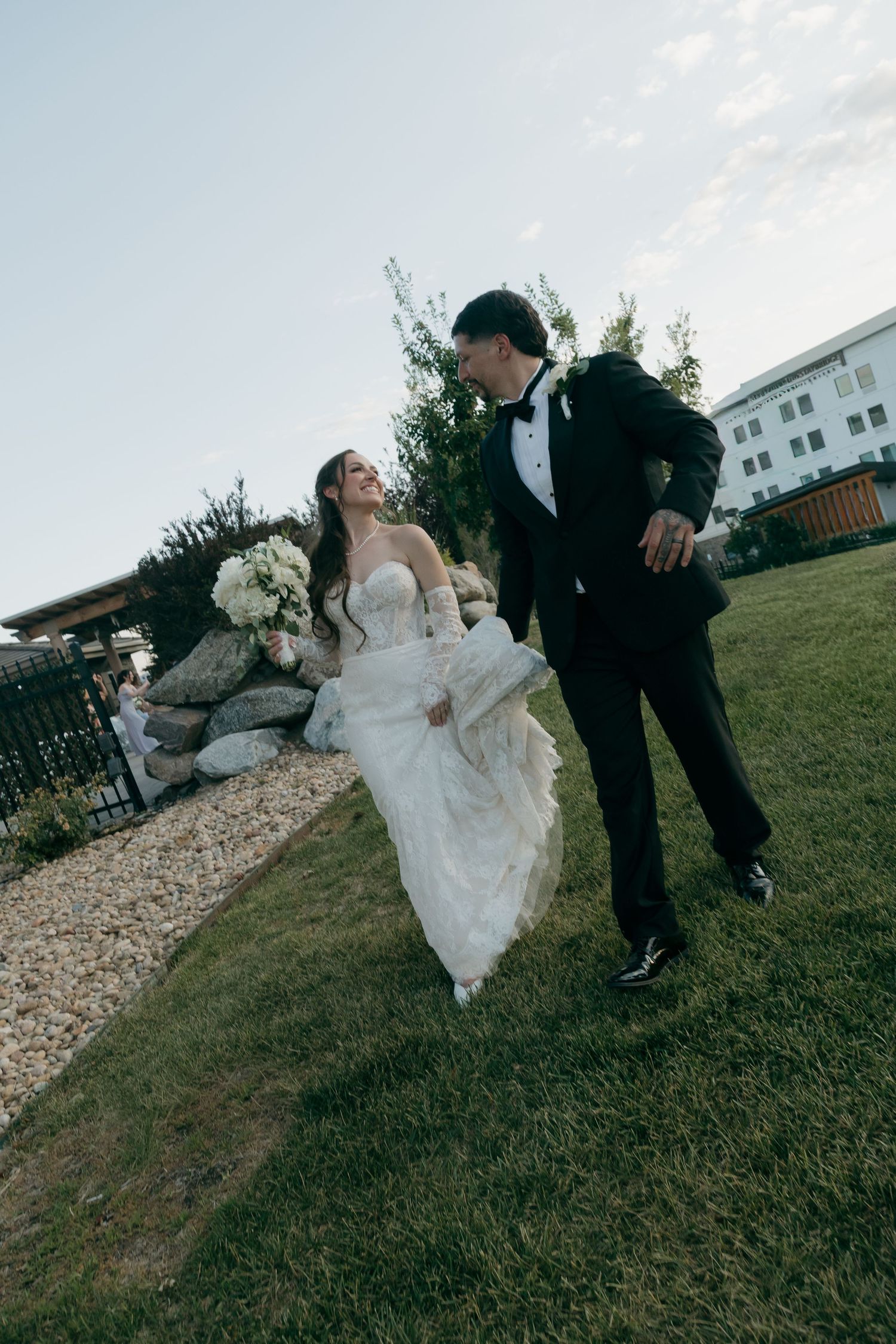 Bride and groom walking together on garden lawn, bride in lace wedding dress holding white bouquet.