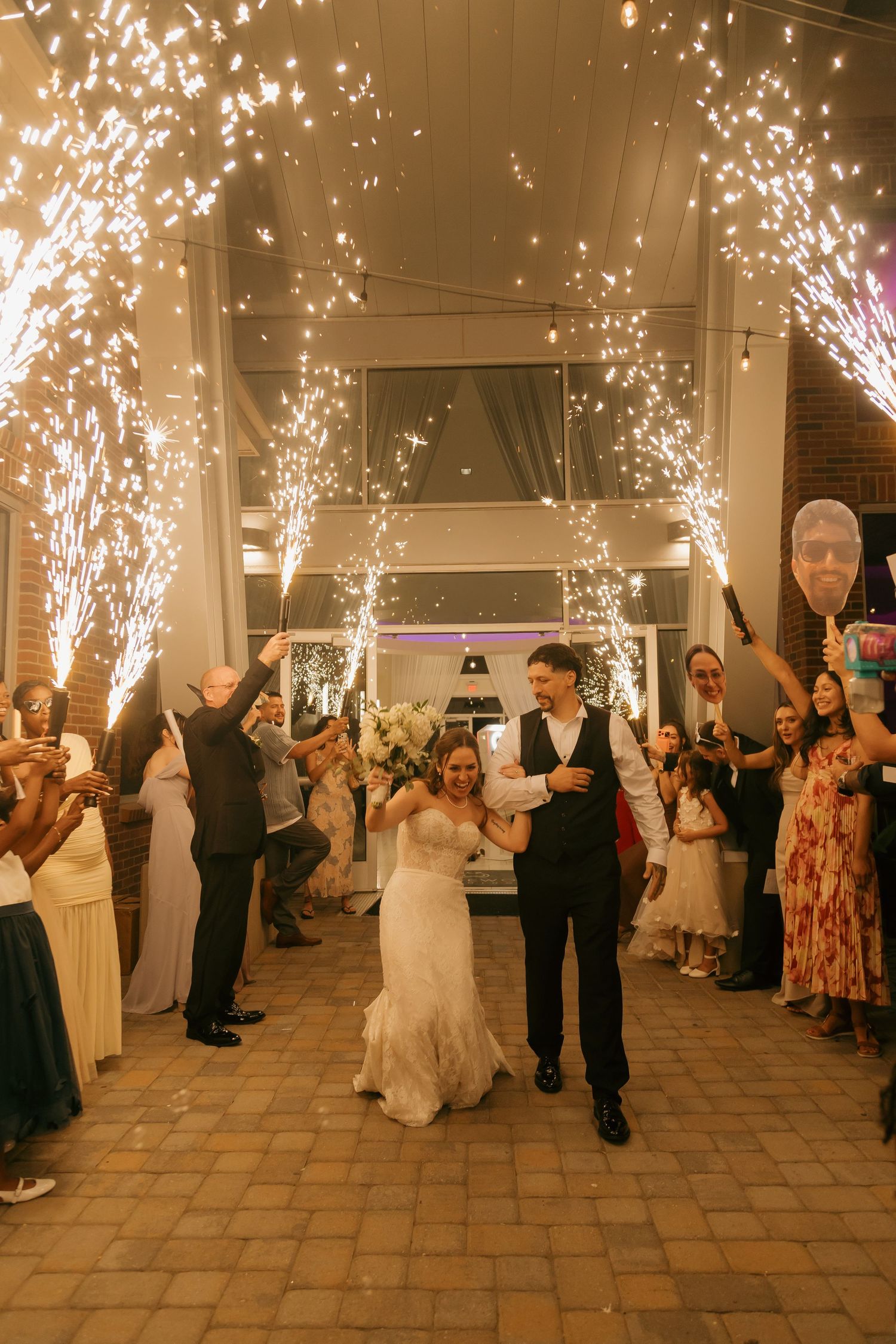 Bride and groom exit wedding reception through dramatic cold spark fountain tunnel with cheering guests.
