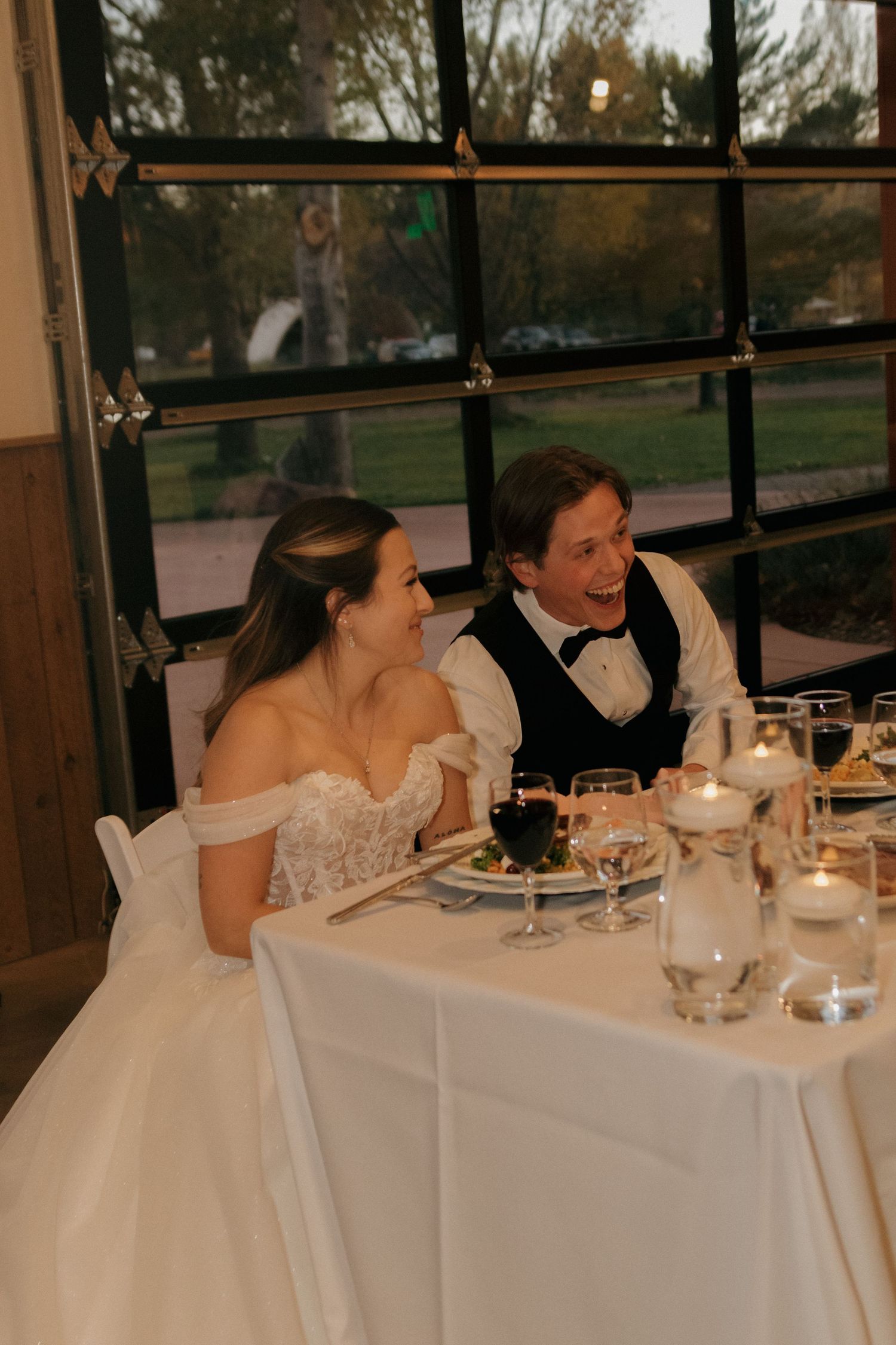 A newlywed couple shares a joyful laugh while seated at their reception dinner table.