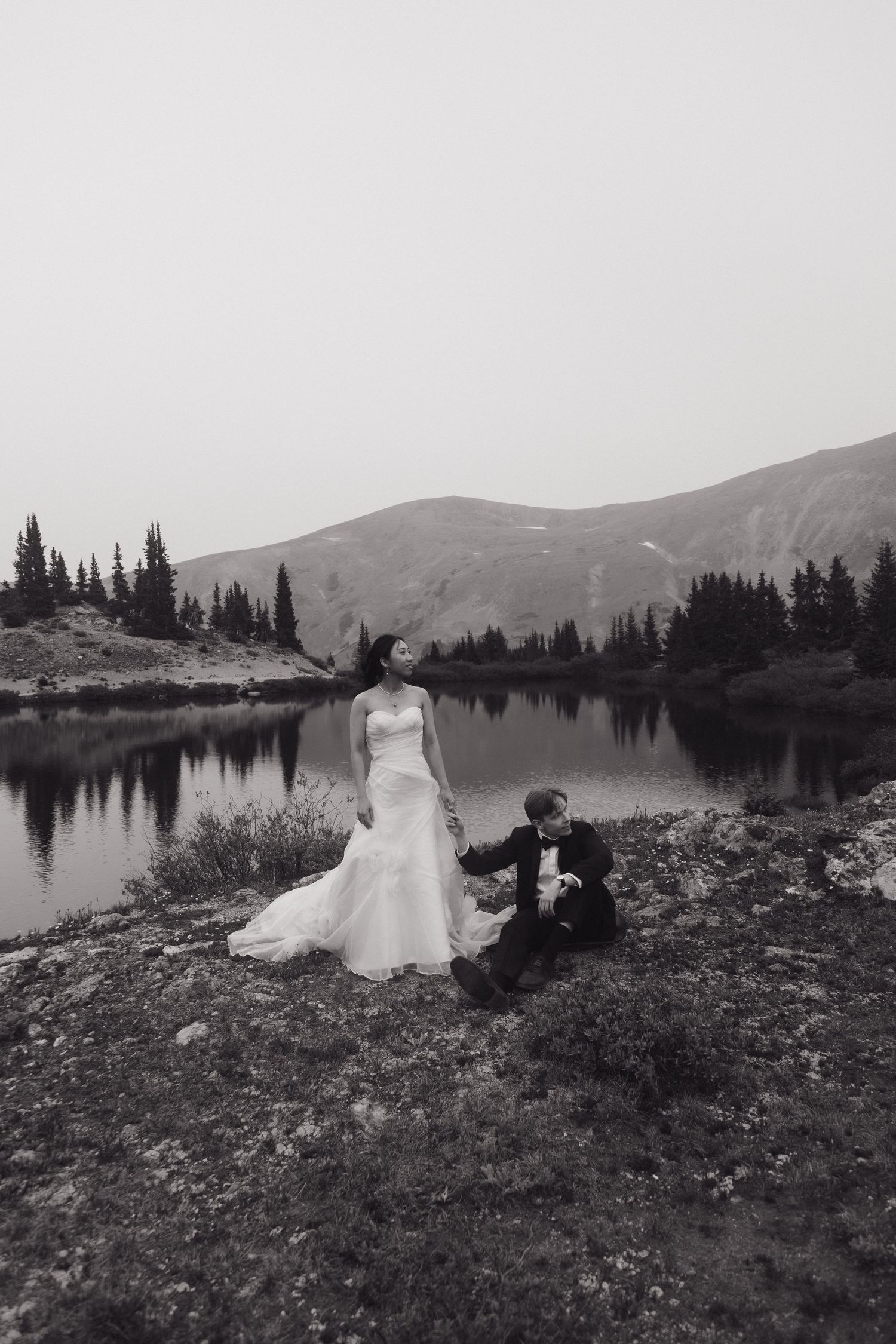 Black and white image of bride standing and groom seated beside a reflective alpine lake at elopement.
