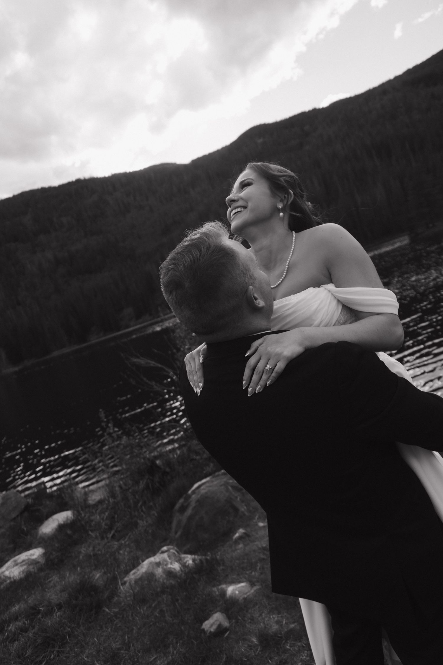 Black and white photo of groom lifting smiling bride by a mountain lake during their wedding celebration.
