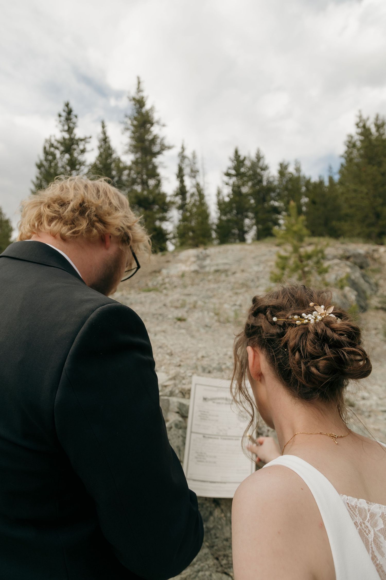 Bride and groom review wedding ceremony documents together outdoors near rocky mountain forest.