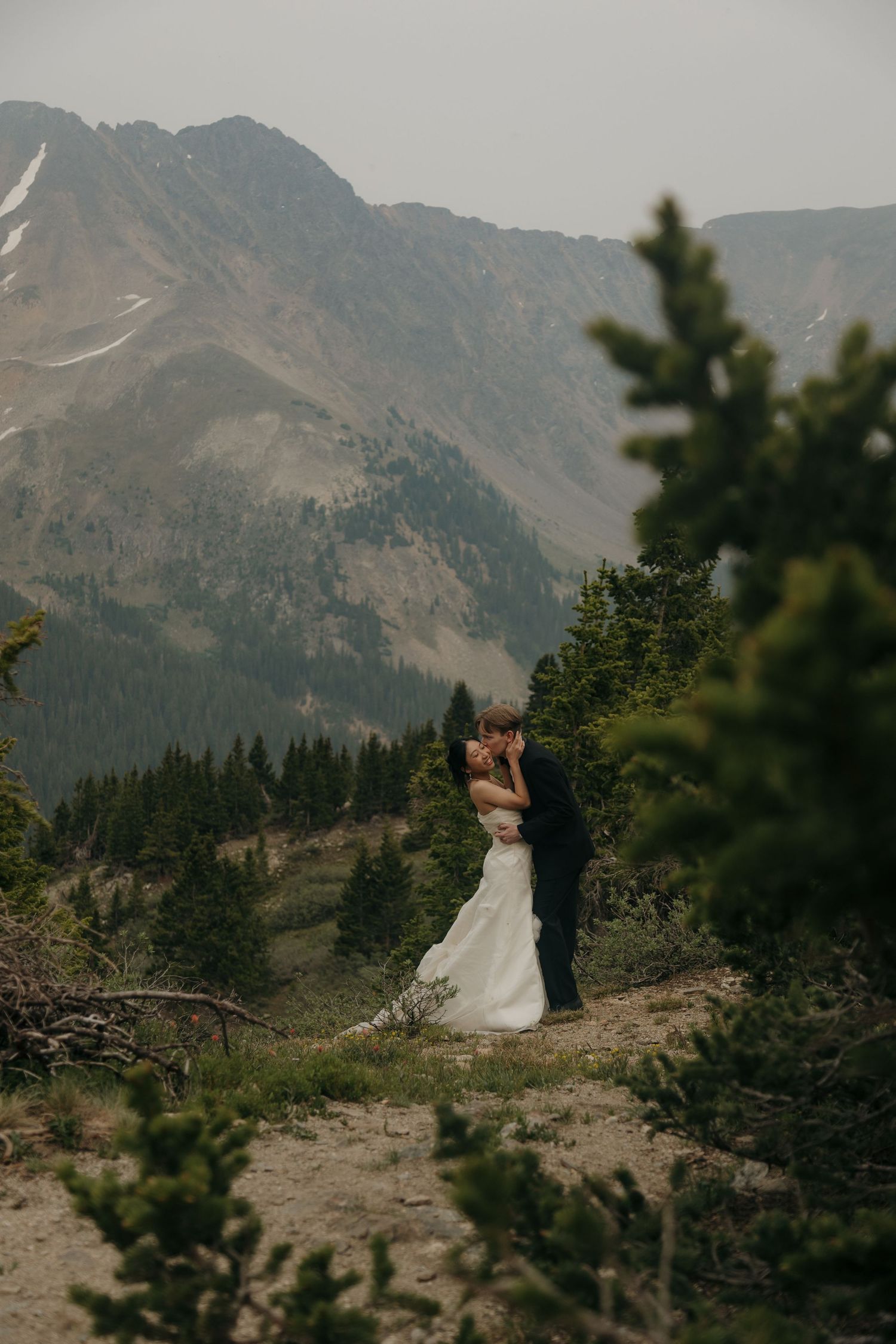 A bride and groom share a romantic kiss on a mountain overlook surrounded by evergreen trees and misty peaks.