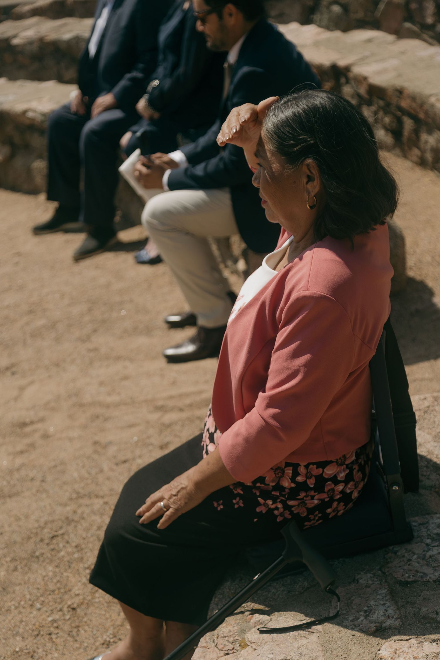 A woman in a pink cardigan shields her eyes from the sun while seated outdoors at a wedding ceremony.