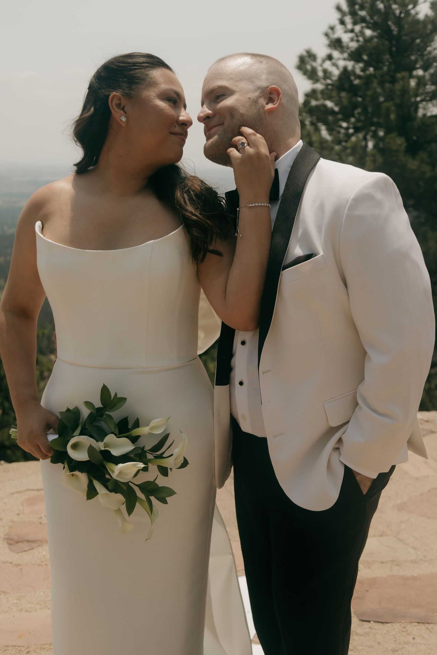A bride and groom in elegant white wedding attire share an intimate moment on a mountain overlook with pine trees.