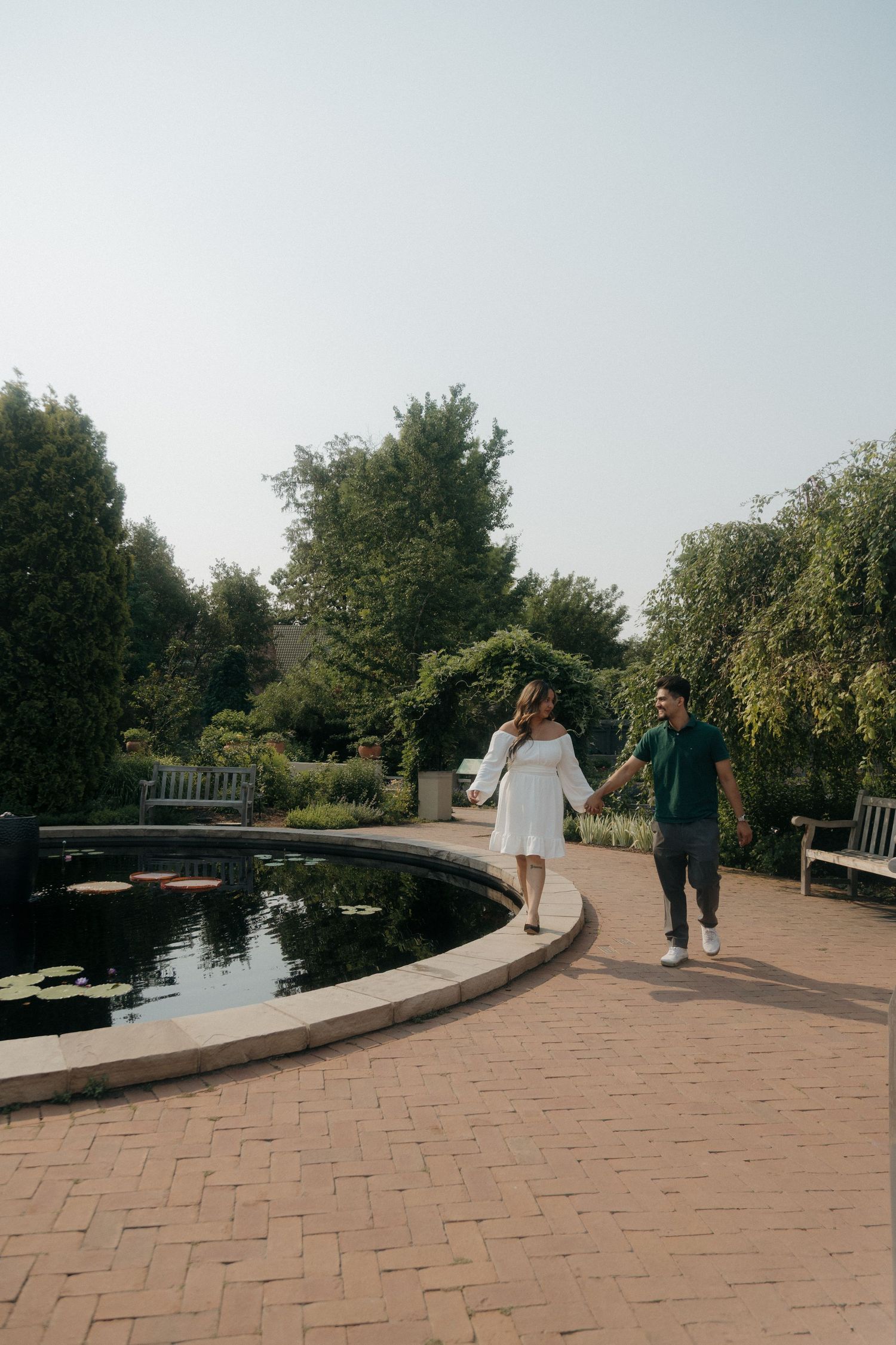 Couple holding hands walking beside a tranquil garden pond surrounded by lush green trees.