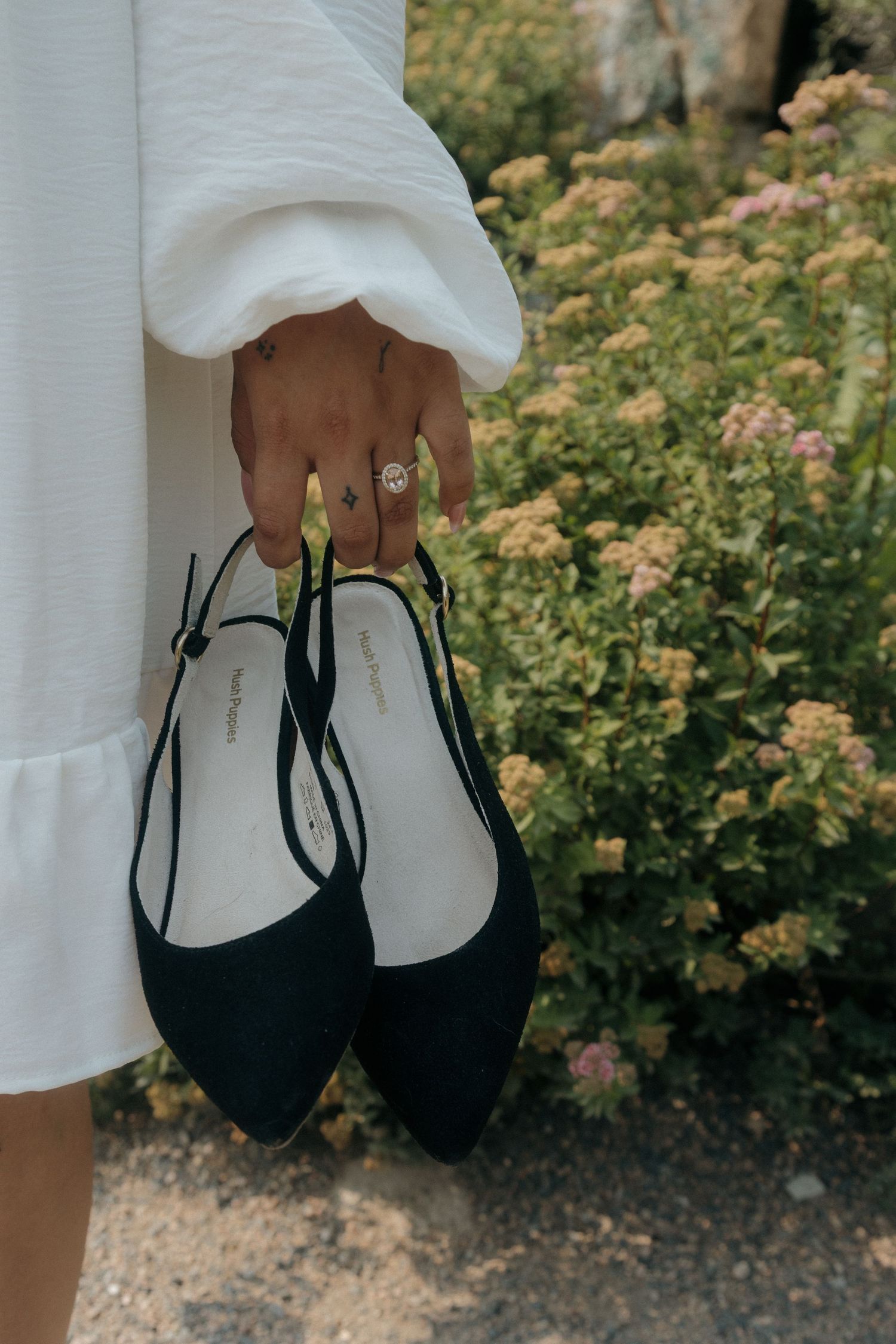 Close-up of a woman holding black slingback heels with a diamond ring visible on her finger near flowers.