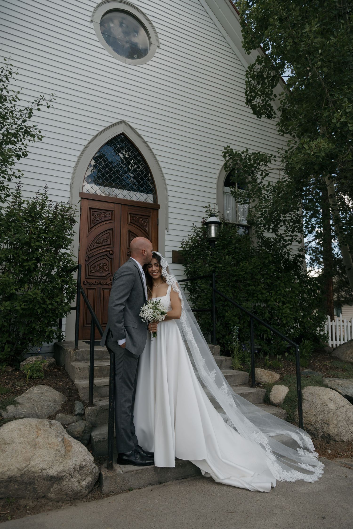 Bride and groom share a romantic moment on the steps of a charming white church with ornate wooden doors.