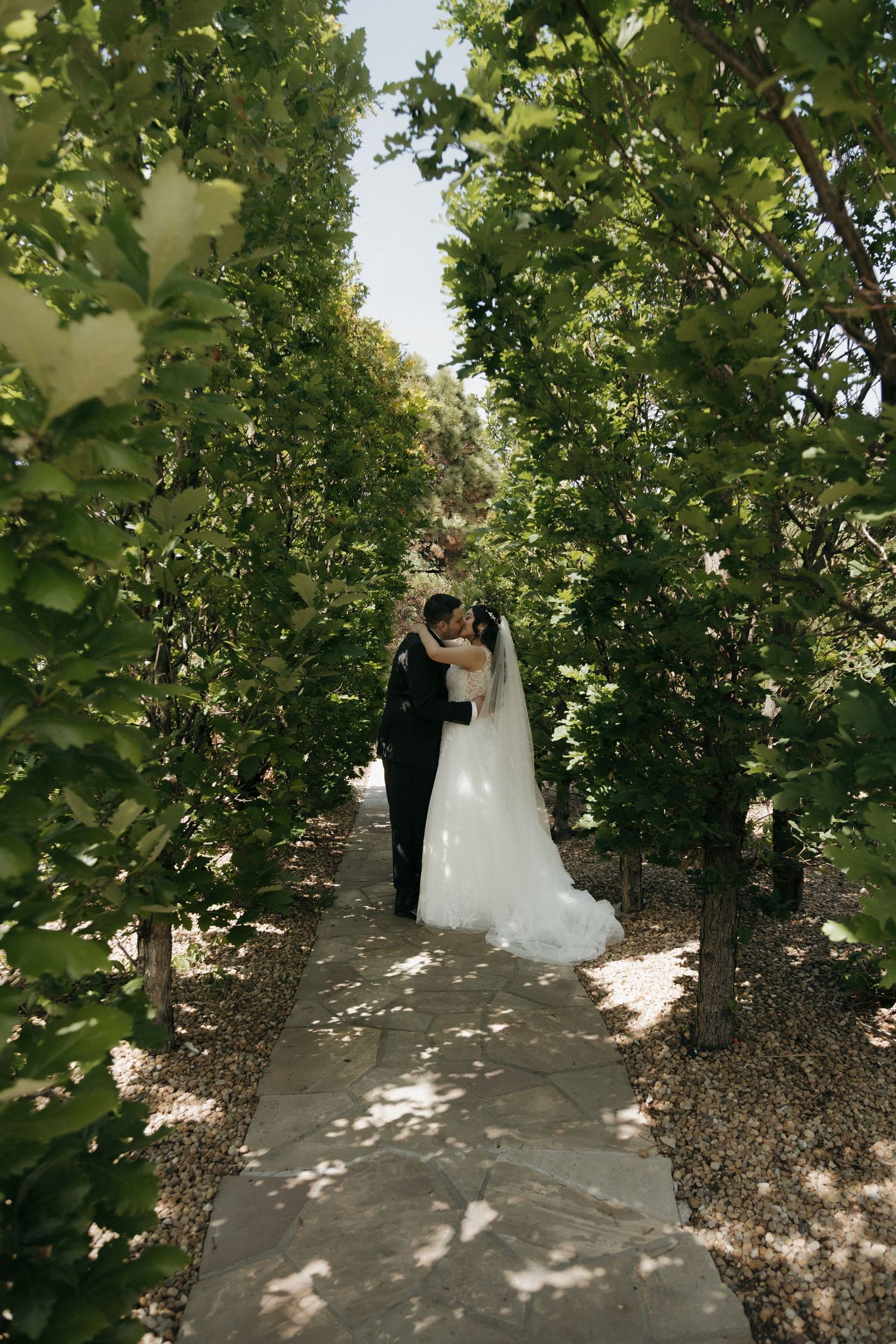 Bride and groom sharing a kiss along a tree-lined garden pathway on their wedding day.