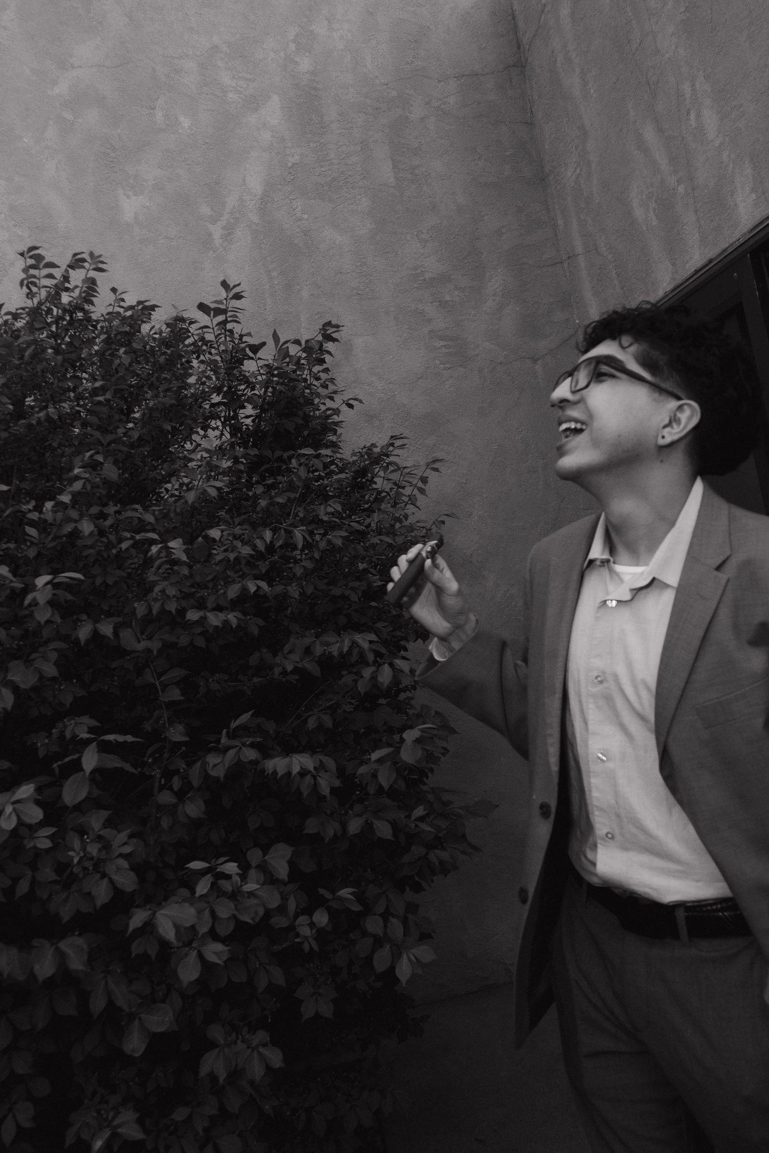 Stylish young man in suit smiles beside lush bush against textured wall in black and white photo.