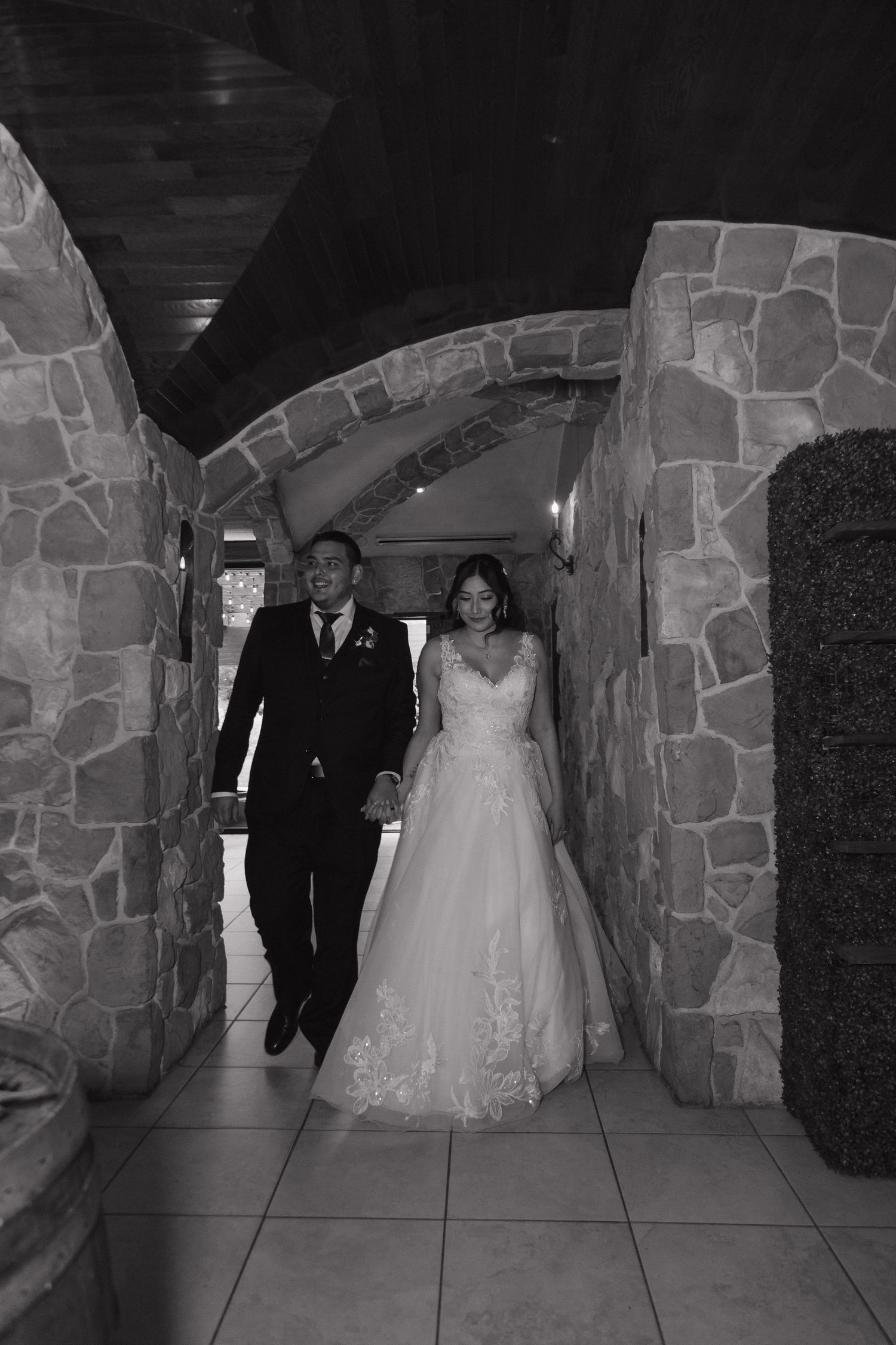 Bride and groom walk hand in hand through stone archway in black and white photo.