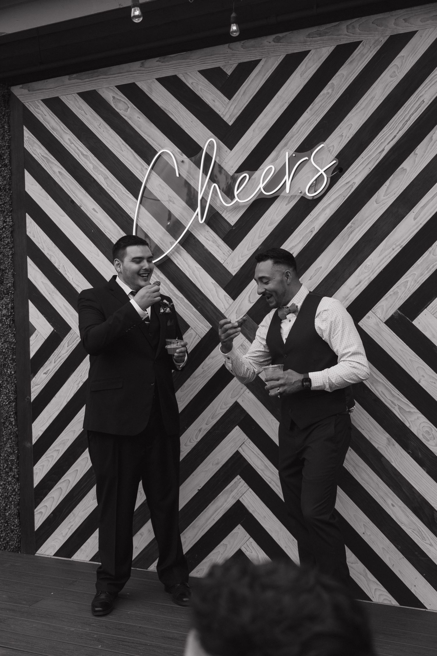 Two men in formal attire toast drinks in front of a Cheers neon sign backdrop.