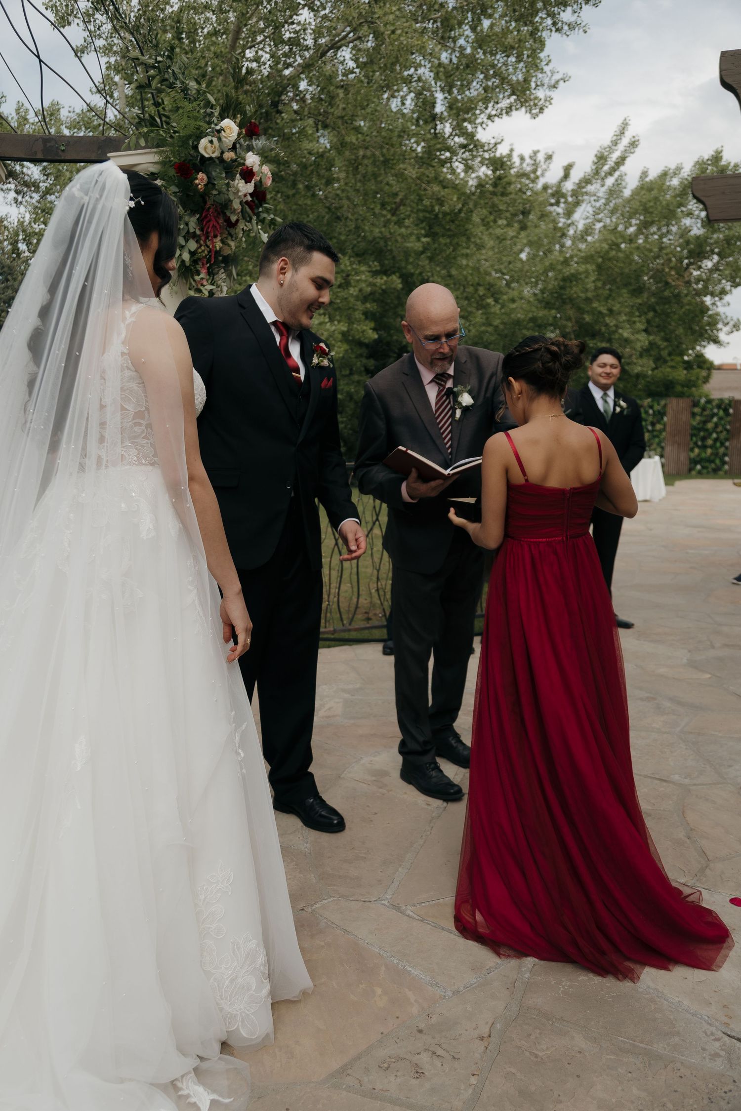 Bride and groom hold hands during outdoor wedding ceremony with officiant and bridesmaid in red gown.