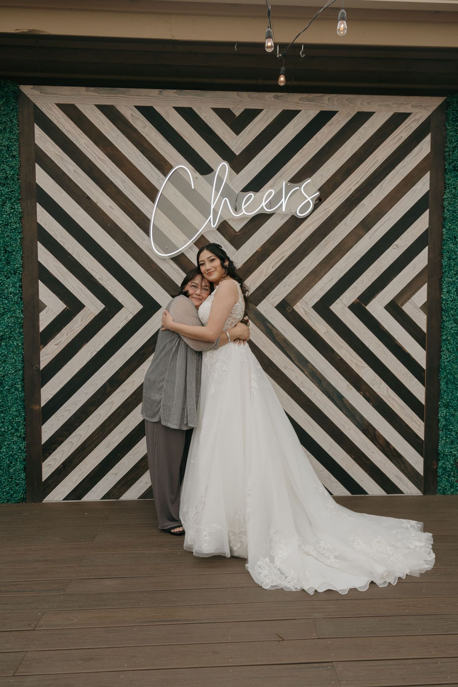 Bride in lace wedding gown embraces a guest in front of a geometric chevron Cheers backdrop at wedding reception.