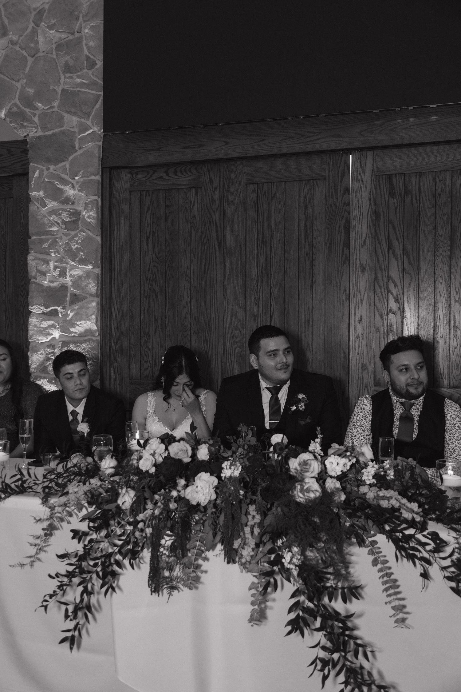 Black and white photo of bride and groom seated at floral wedding head table with guests.