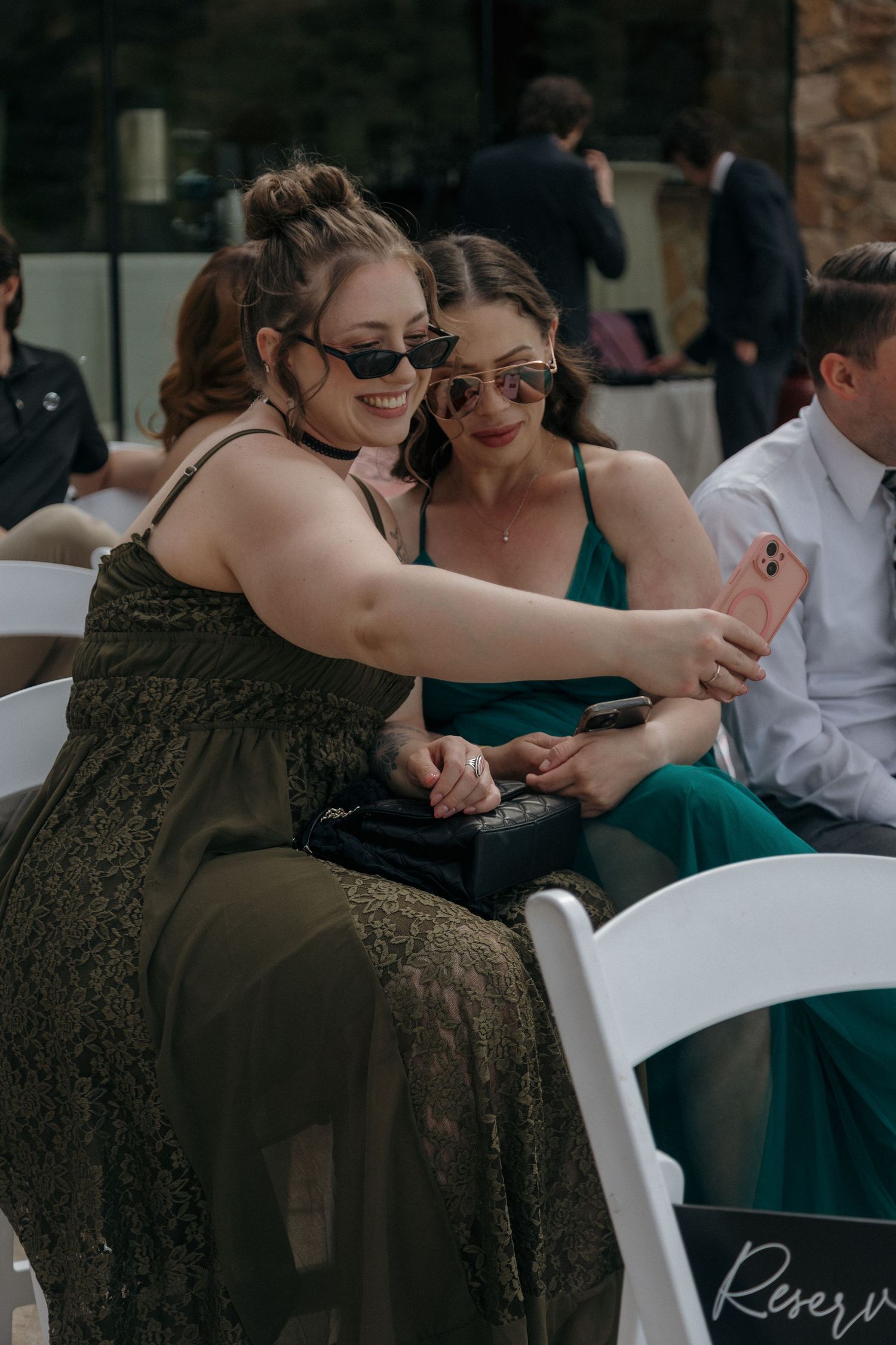 Two wedding guests in sunglasses take a selfie while seated at an outdoor ceremony.