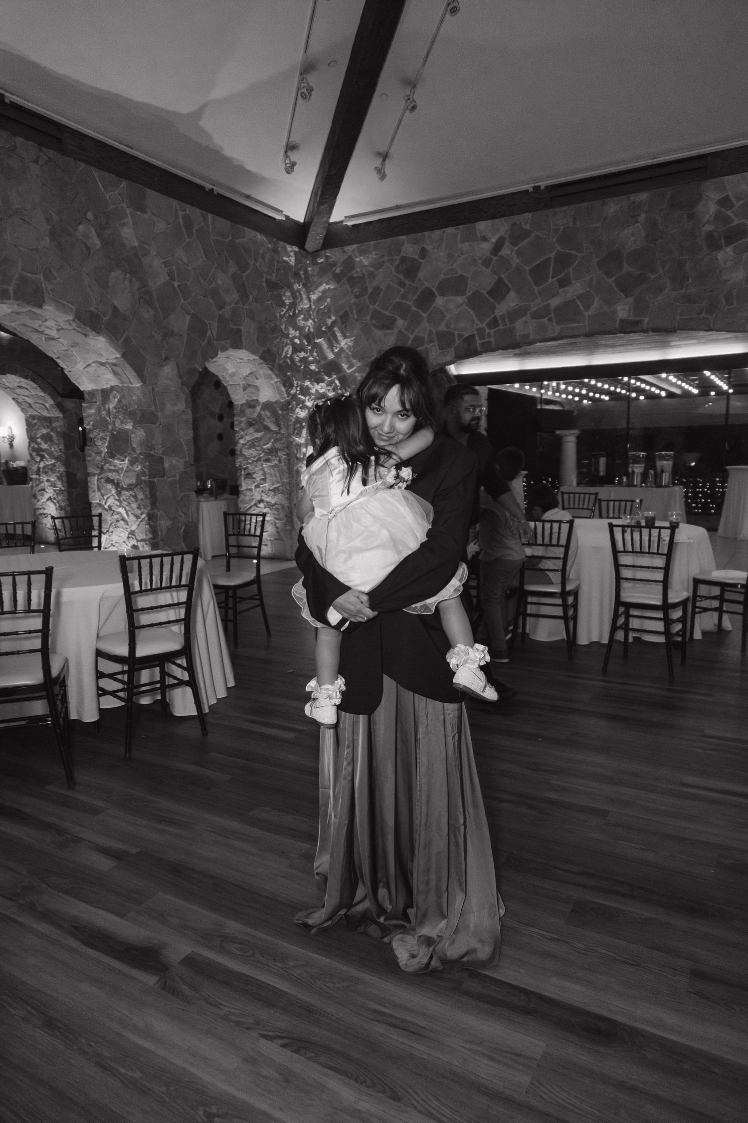 Black and white photo of woman in formal gown carrying baby at wedding reception venue.