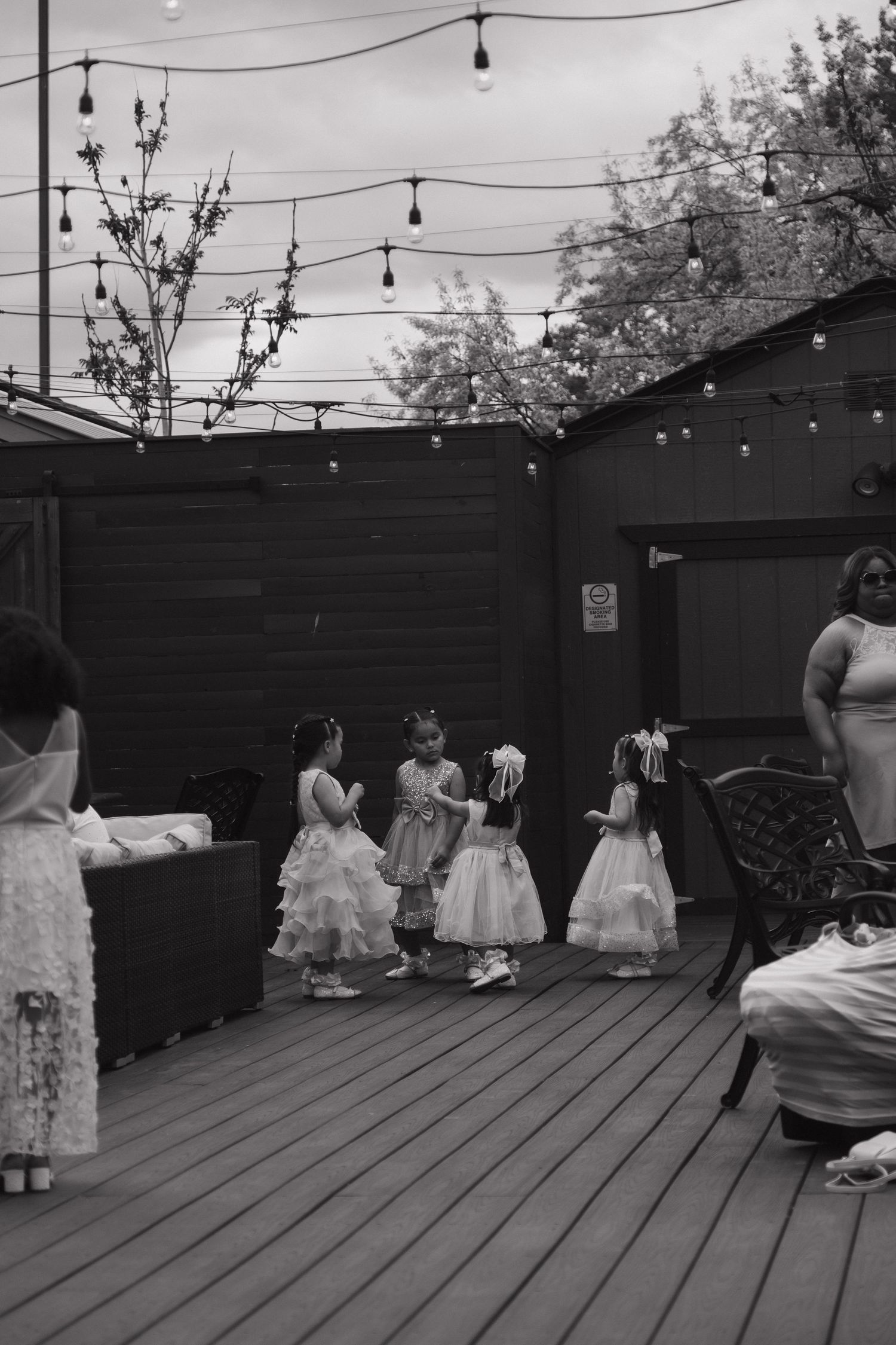 Young flower girls in white dresses gather on decorated outdoor deck at wedding celebration.
